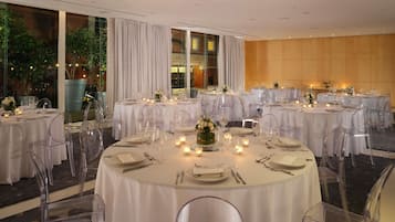 A banquet hall with round tables set for dining, clear chairs, and candles on each table.