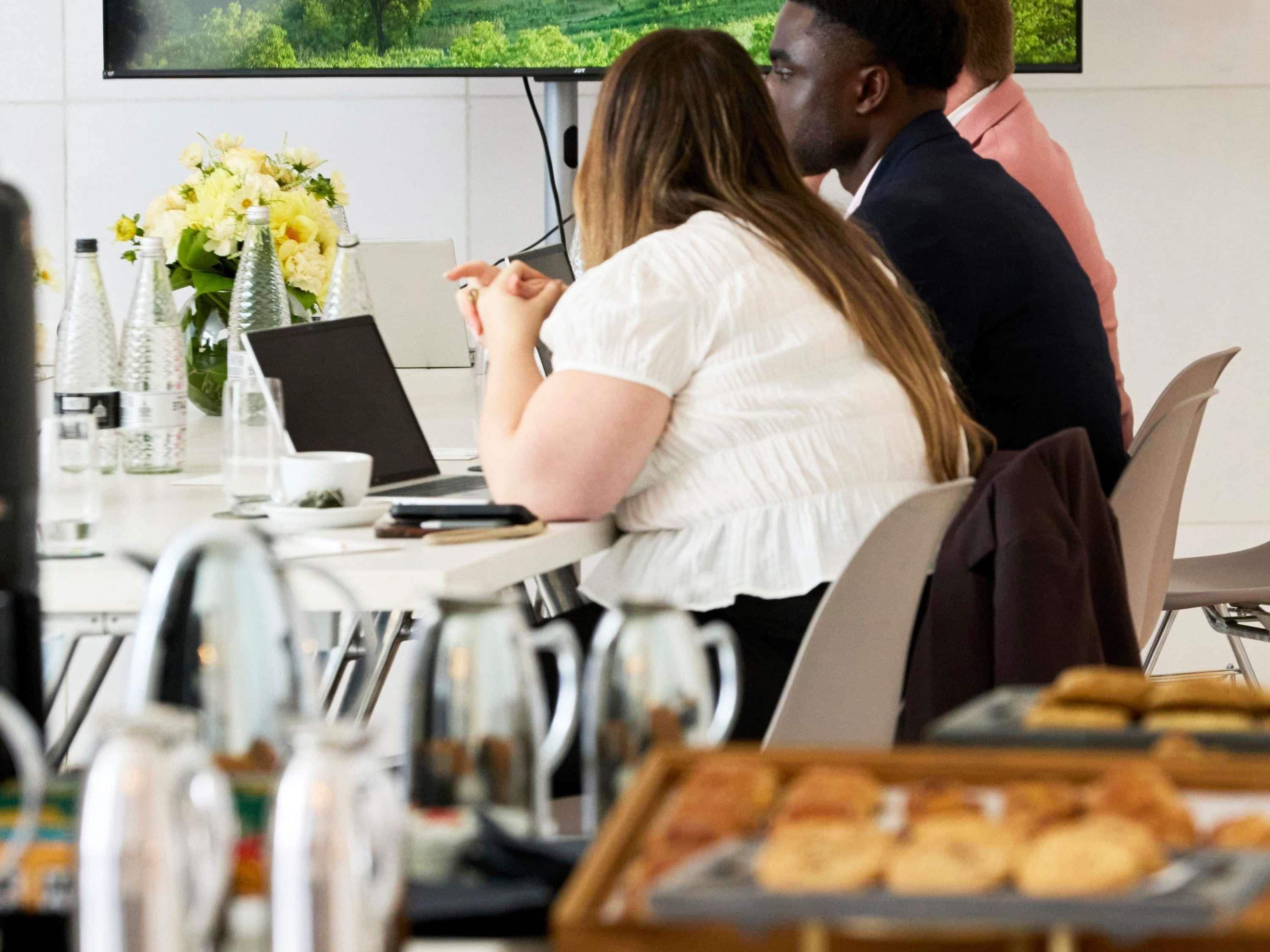 Une salle de conférence avec un ordinateur portable, un bouquet de fleurs et un plateau de pâtisseries sur la table.