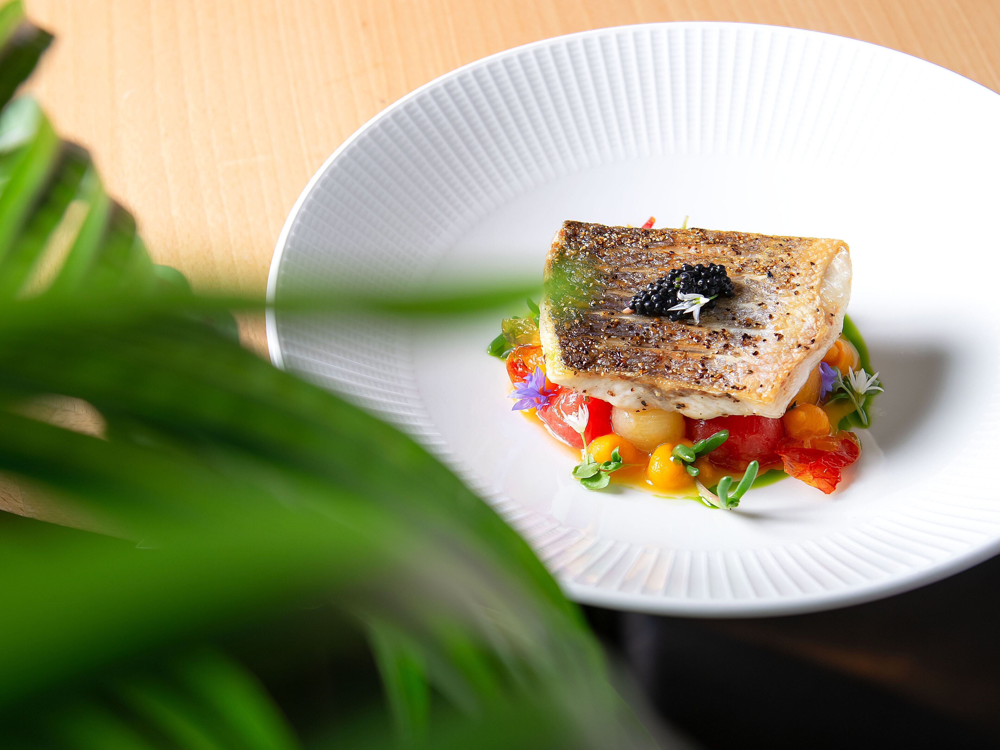 A white plate with a piece of fish, garnished with black caviar and surrounded by colorful vegetables.