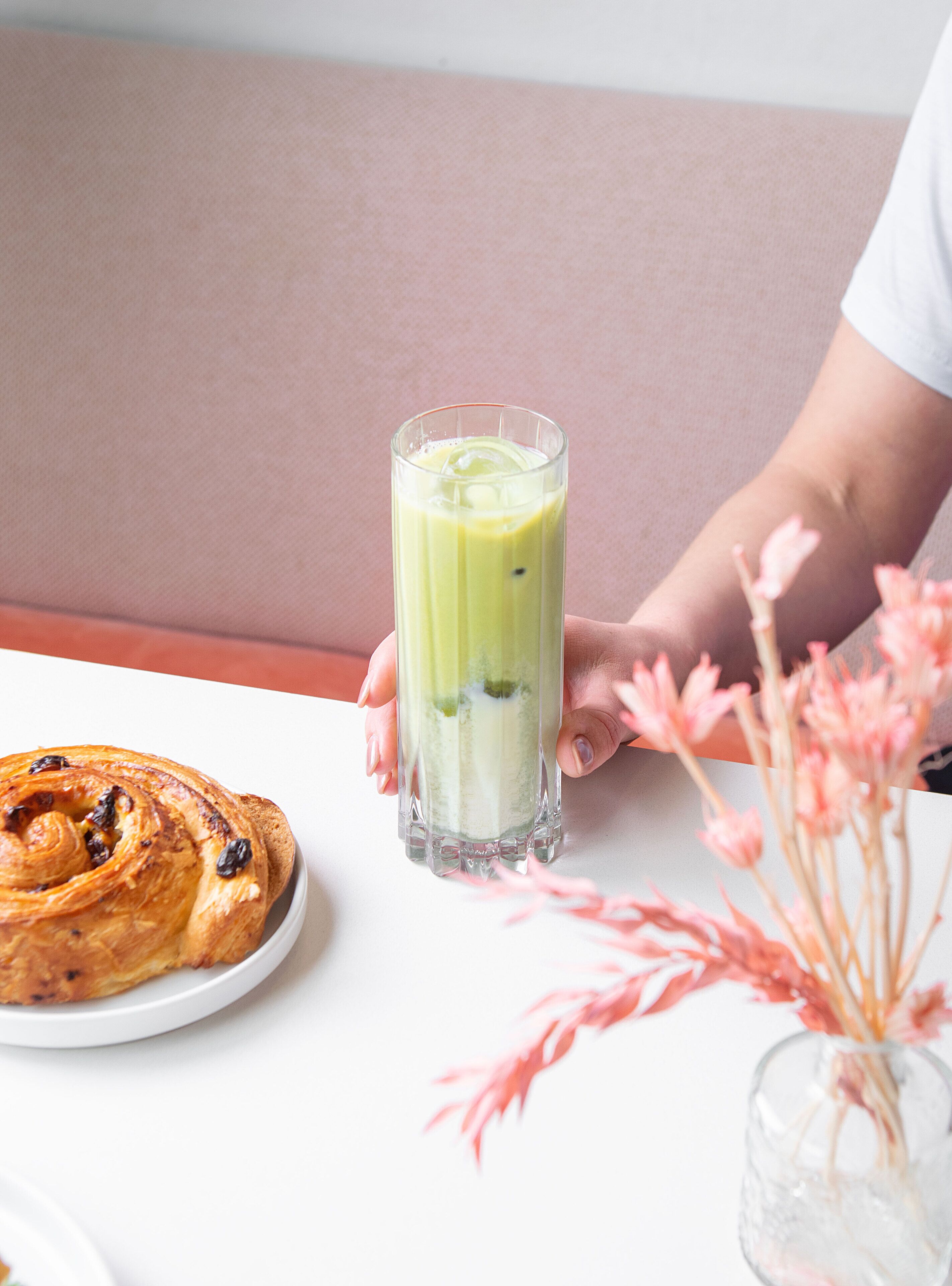A person holding a tall glass of green smoothie, a pastry on a plate, and a vase with pink flowers on a table.