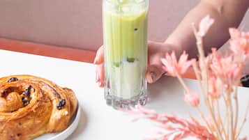 A person holding a tall glass of green smoothie, a pastry on a plate, and a vase with pink flowers on a table.