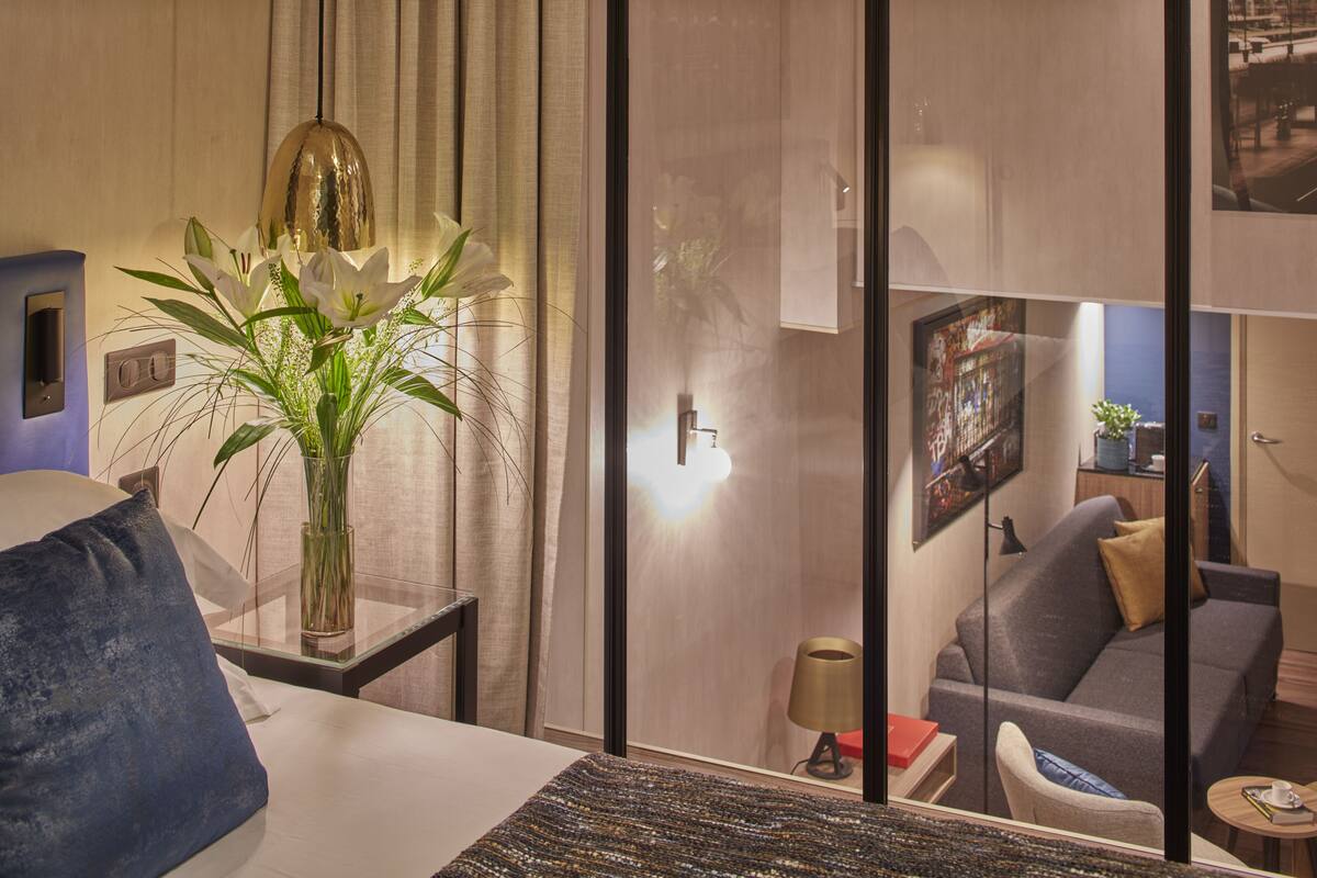 Une chambre d’hôtel moderne avec un grand lit, une table de chevet avec une lampe, un vase rempli de fleurs et une vue sur une autre chambre avec un canapé et un tableau.