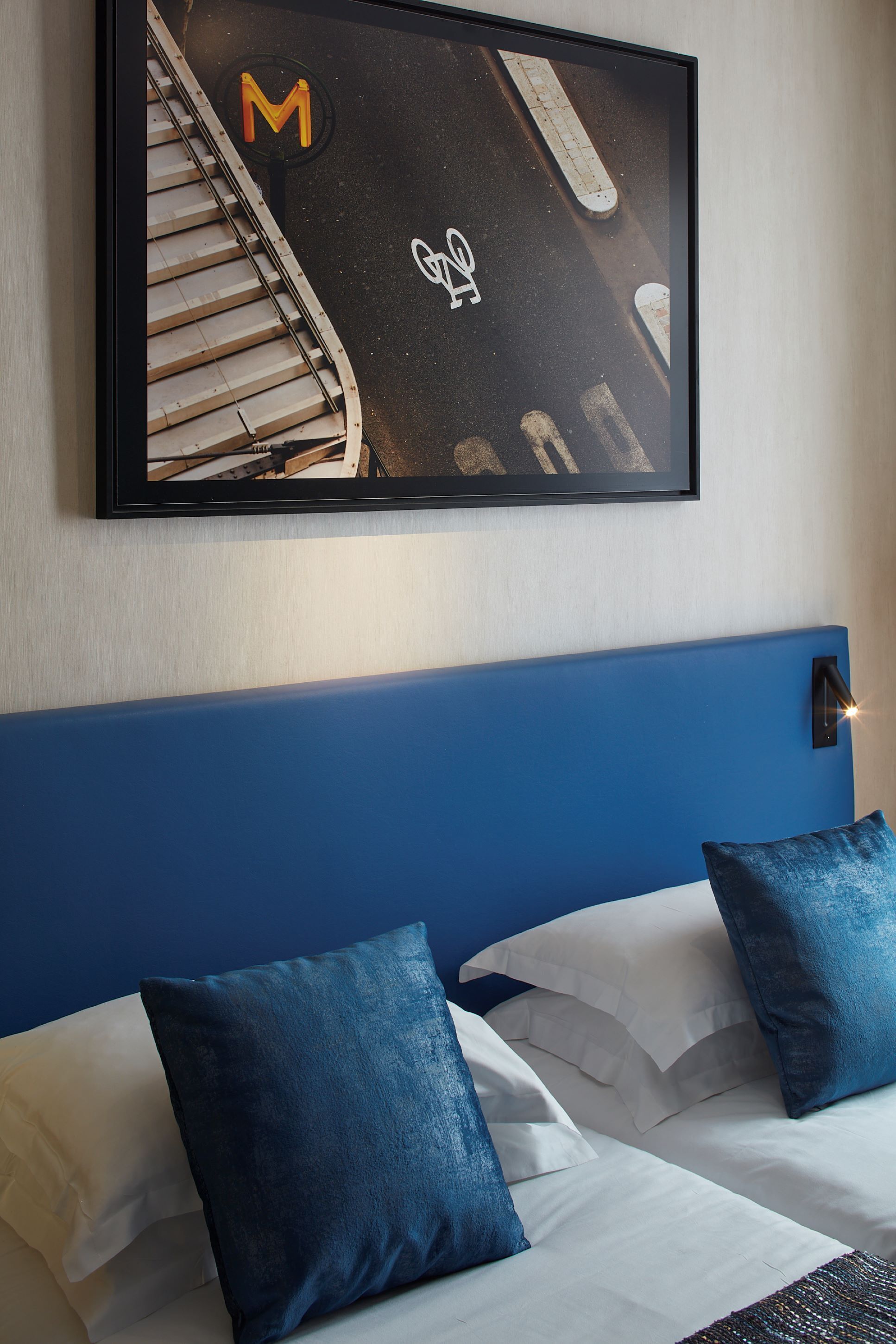 Une chambre d’hôtel avec une tête de lit bleue, du linge de lit blanc et un tableau représentant une scène de rue accroché au mur.