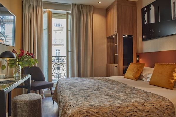 A hotel room with a bed, a desk with a lamp, a chair, a potted plant, and a view of the outside.