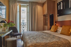 Habitación de hotel con cama, escritorio con lámpara, una silla, planta en maceta y vistas al exterior.