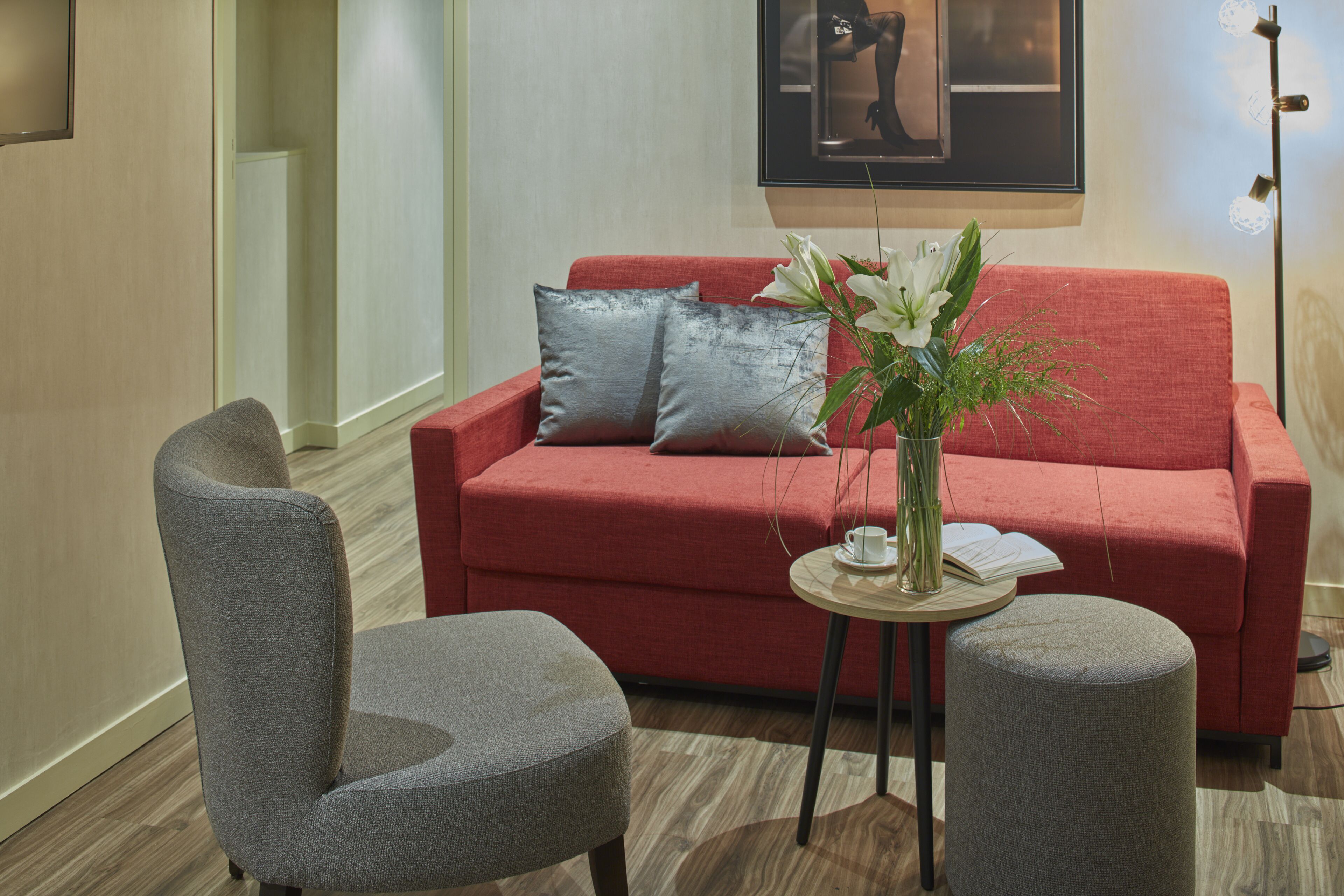 Ein modernes Wohnzimmer mit einem roten Sofa, zwei grauen Sesseln, einem kleinen runden Tisch mit einem Blumenstrauß und einem gerahmten Bild an der Wand.