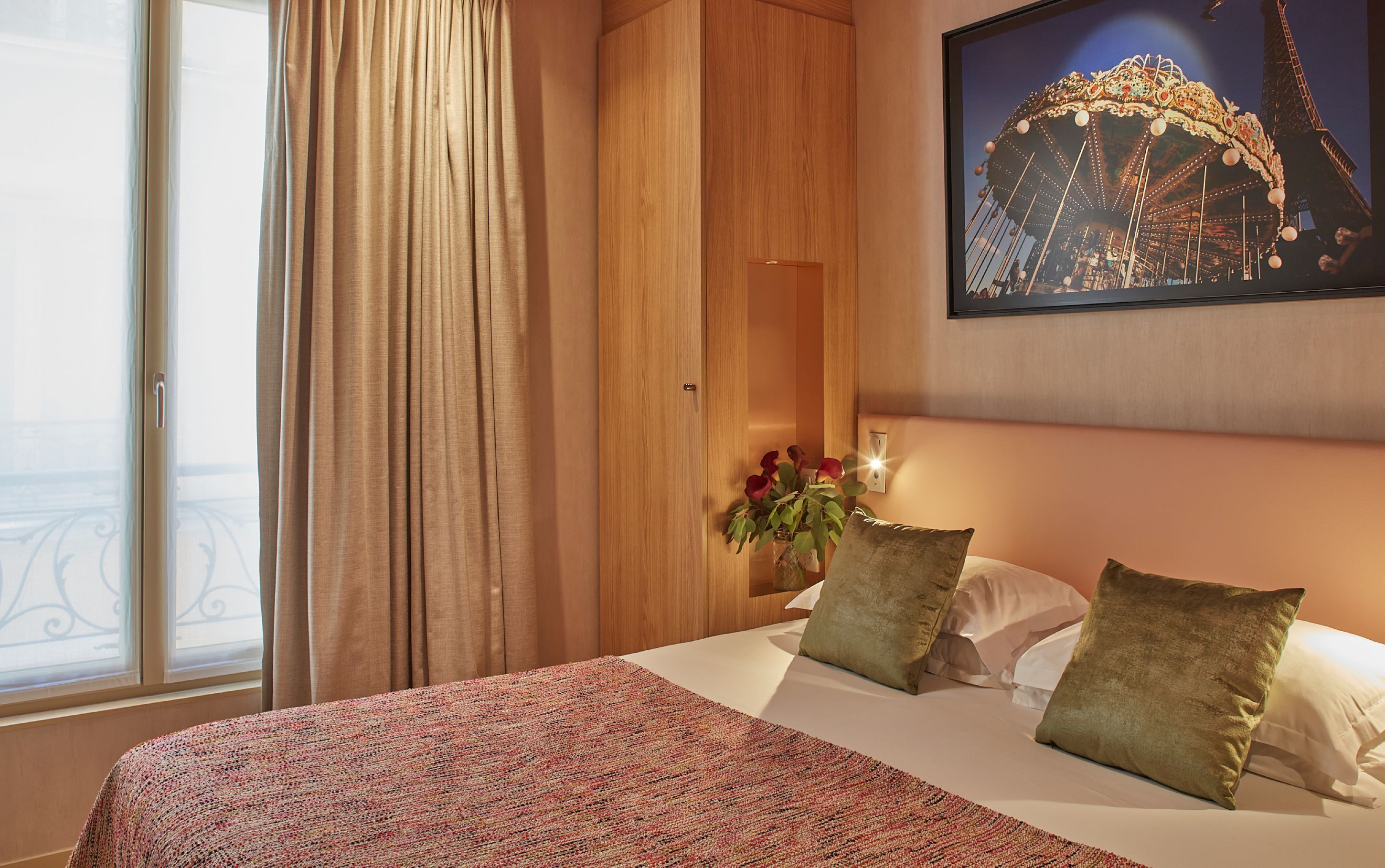 A hotel room with a bed, a window with curtains, a wooden wardrobe, a wall-mounted picture of a Ferris wheel, and a vase with flowers.