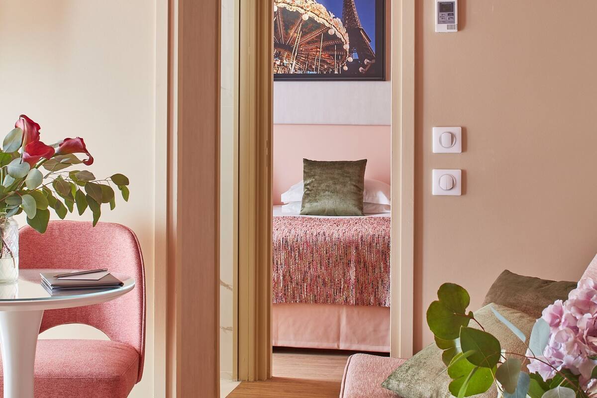 Une chambre d’hôtel moderne avec un canapé rose, une petite table et une vue sur une grande roue, visible à travers une porte ouverte.