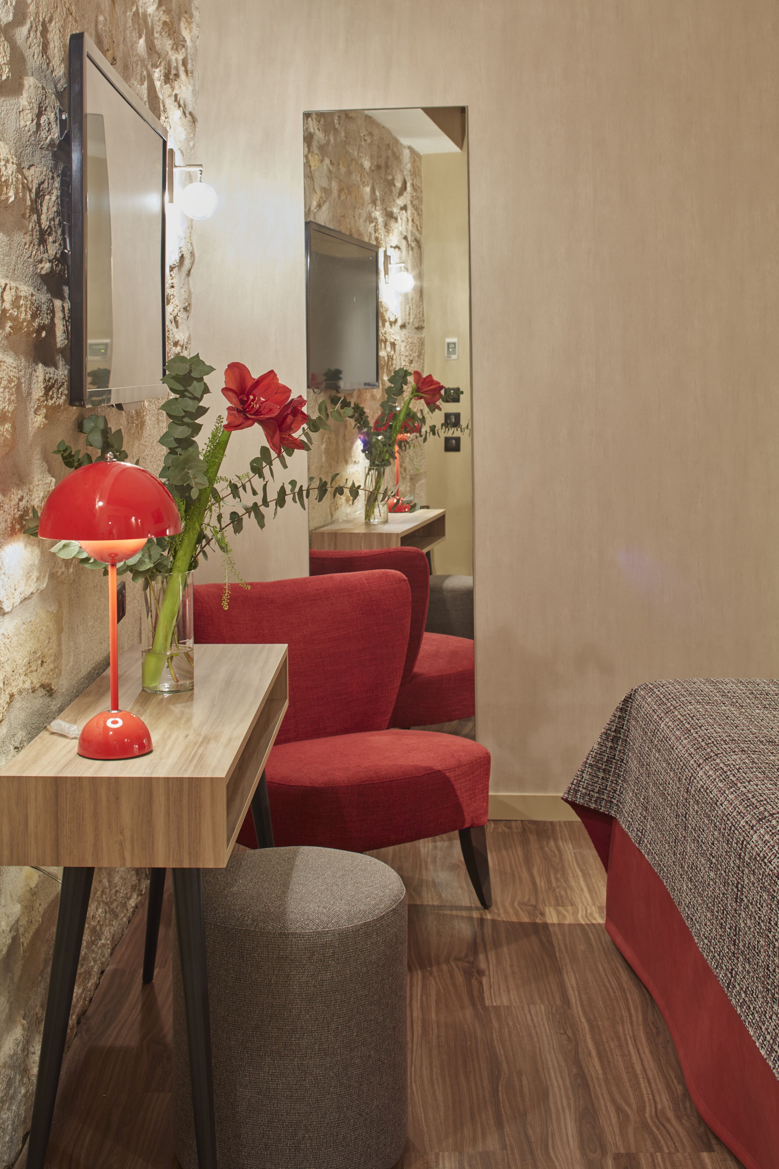 Una habitación de hotel moderna con un escritorio de madera, una silla roja y una lámpara roja. Hay un espejo en la pared y un taburete pequeño y redondo.