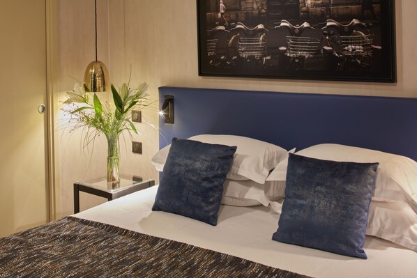A hotel room with a bed, two pillows, a nightstand with a lamp, a vase with flowers, and a framed picture on the wall.