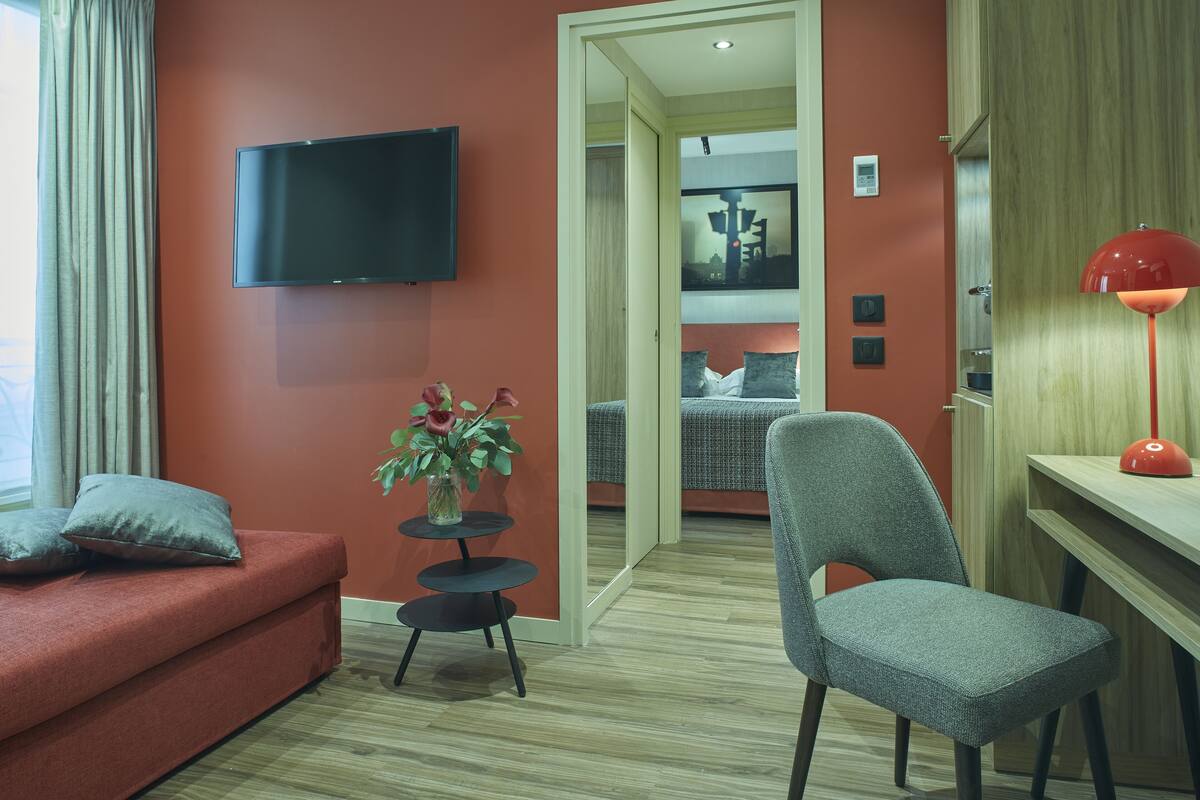 Une chambre d’hôtel moderne avec un mur rouge, un téléviseur à écran plat fixé au-dessus d’un canapé, une petite table ornée d’un vase de fleurs et un bureau avec une lampe rouge.