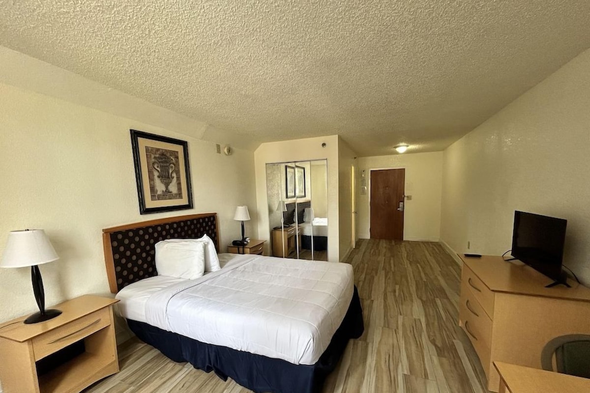 A hotel room with a bed, bedside table, lamp, TV, and a desk.