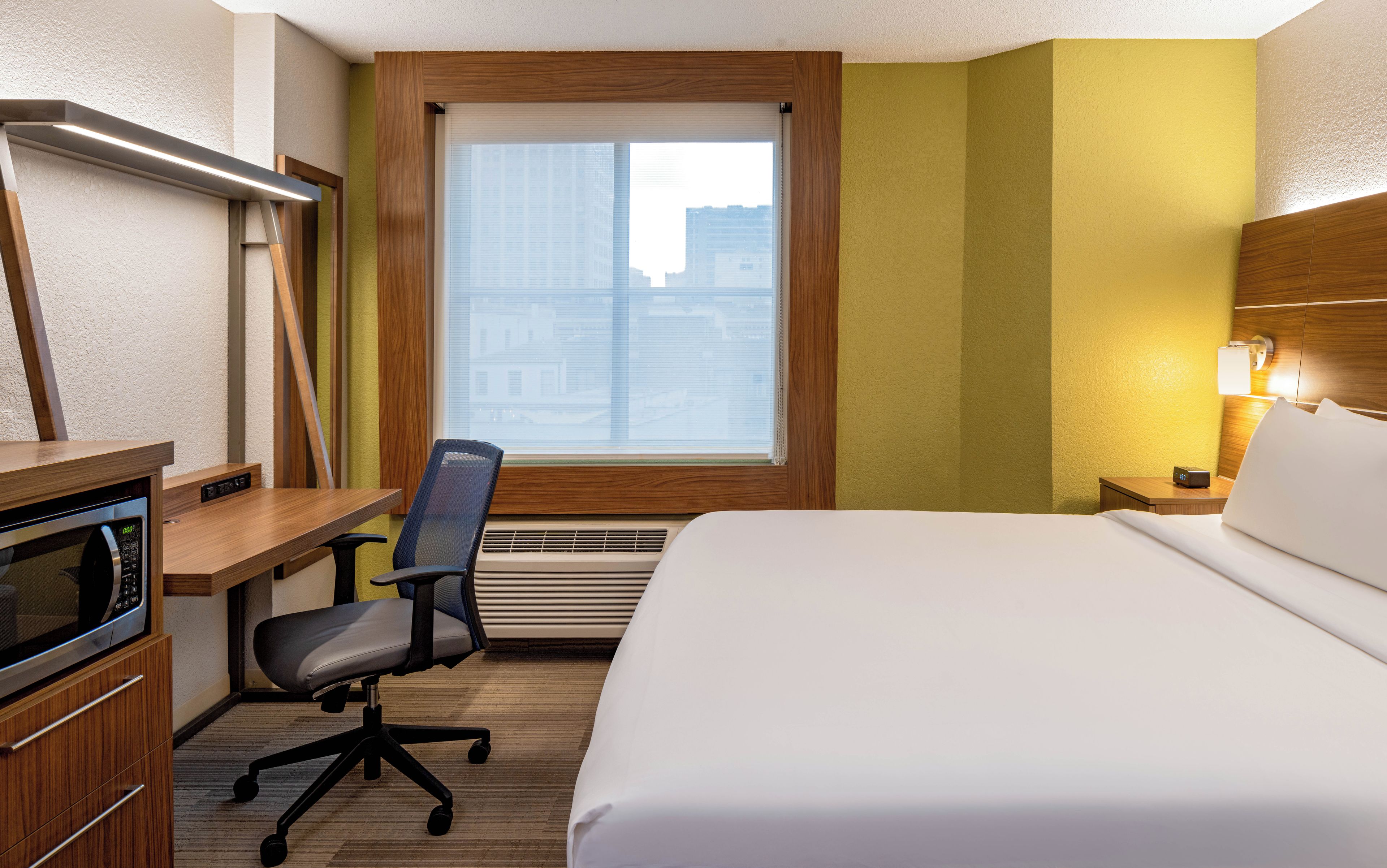 A hotel room with a bed, desk, chair, microwave, and a window with blinds.