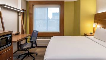 A hotel room with a bed, desk, chair, microwave, and a window with blinds.