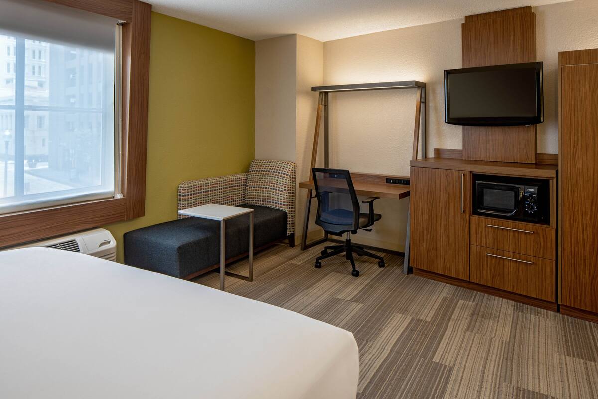 A hotel room with a bed, a desk with a chair, a television, a microwave, and a window with a view of buildings.