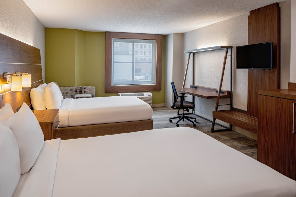 A hotel room with two beds, a desk, a chair, a TV, and a window with a view of buildings.