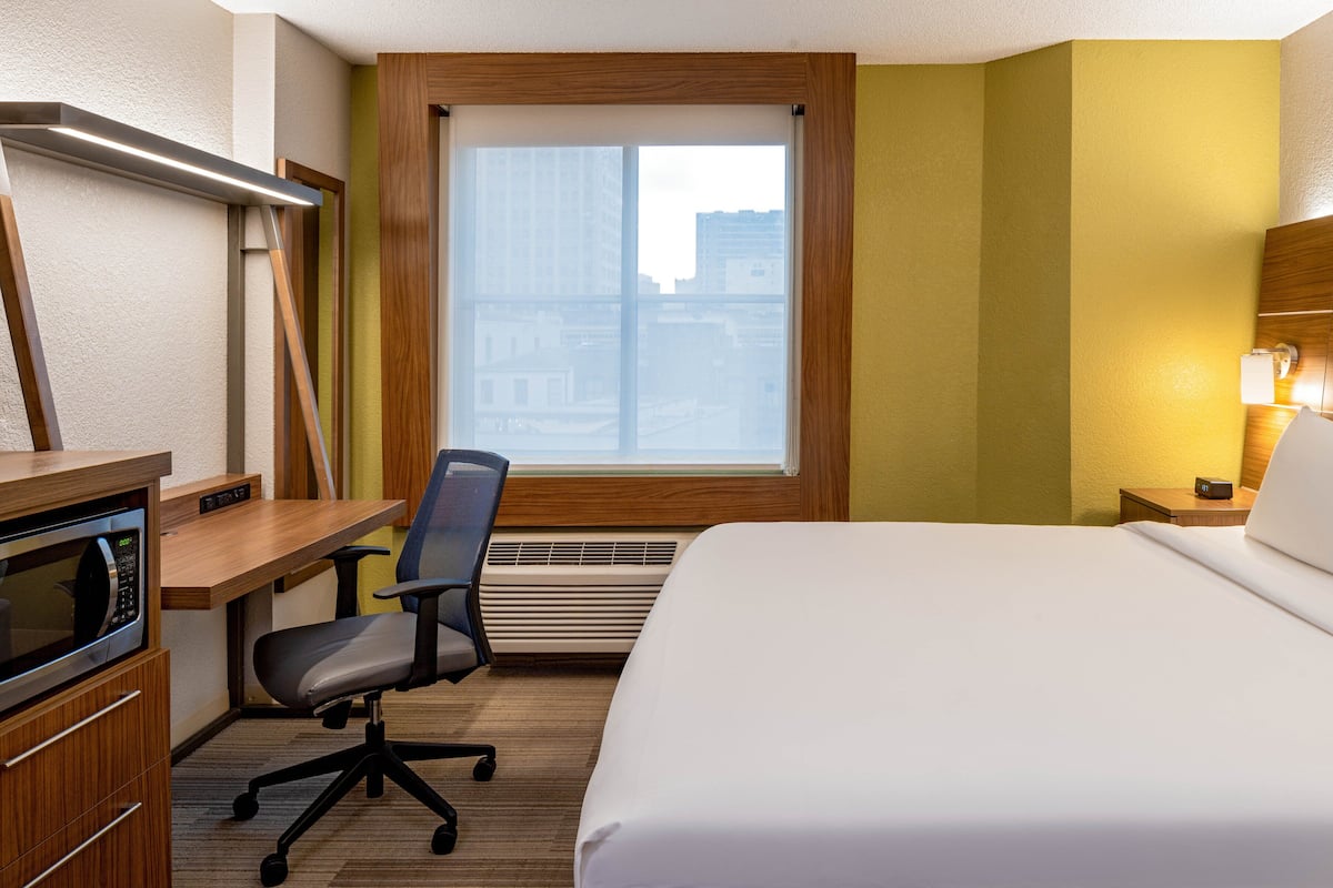 A hotel room with a bed, desk, chair, microwave, and a window with blinds.