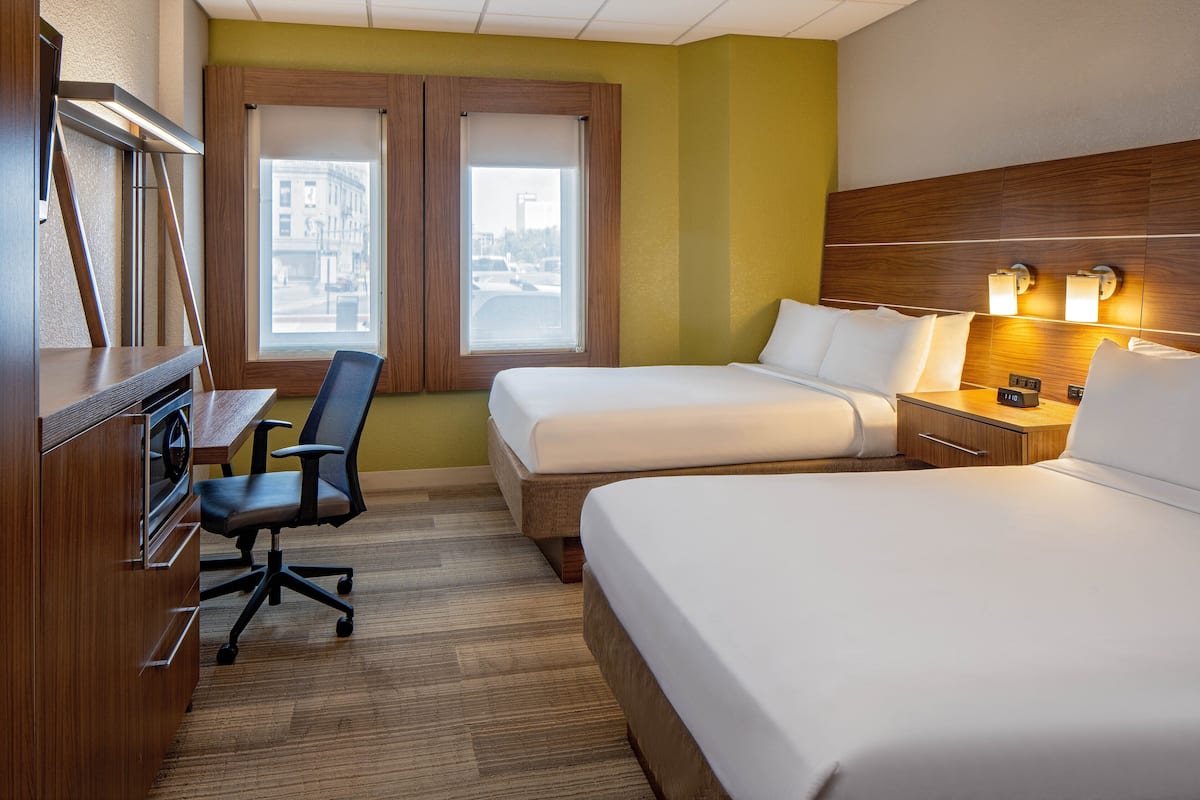 A hotel room with two beds, a desk, and a chair. There are windows with a view of buildings outside.