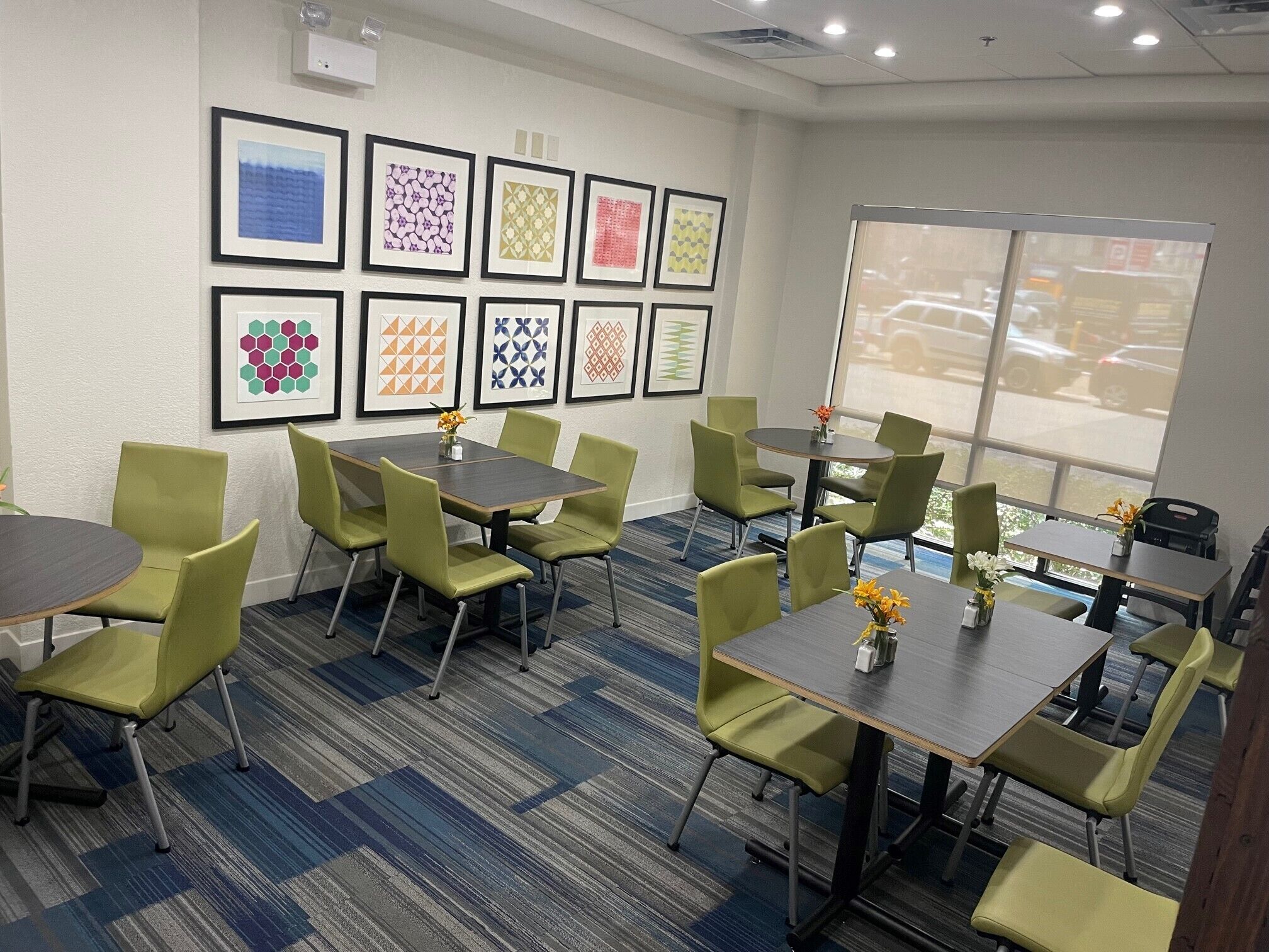 A conference room with green chairs and tables, framed artwork on the wall, and a view of the parking lot through large windows.