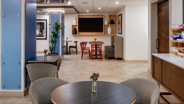 A modern dining area with a round table, grey chairs, and a vase of flowers. There are red chairs and a small table with a plant in the background. - Holiday Inn Express New Orleans Downtown by IHG (New Orleans)