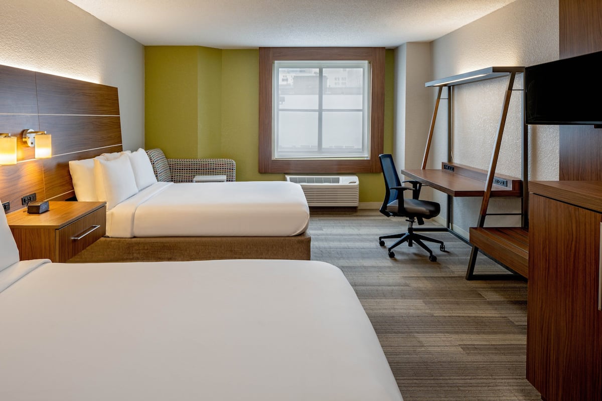 A hotel room with two beds, a desk, a chair, a TV, and a window with a view of buildings.