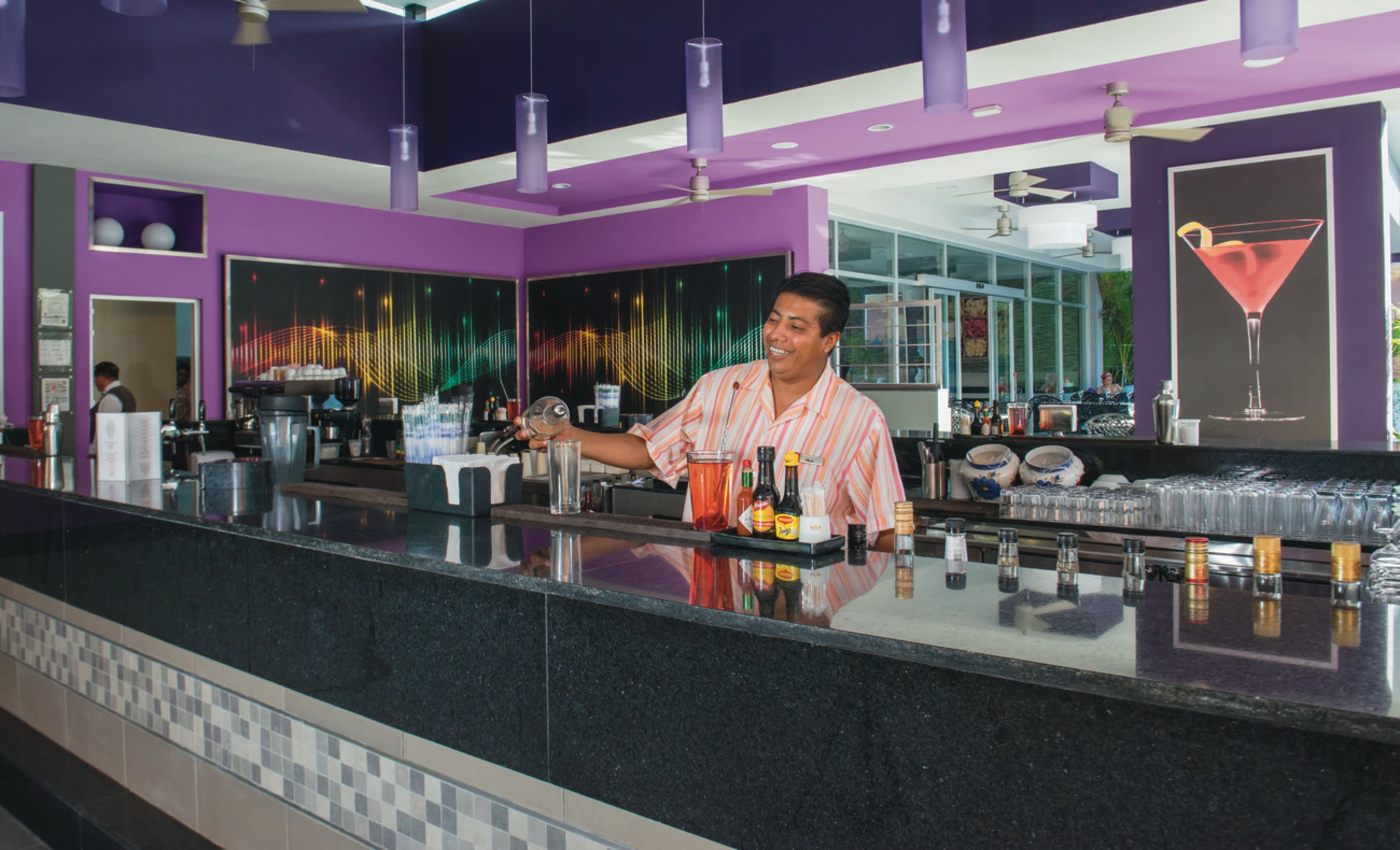 A bar area with a bartender, purple decor, and a martini glass on the wall.