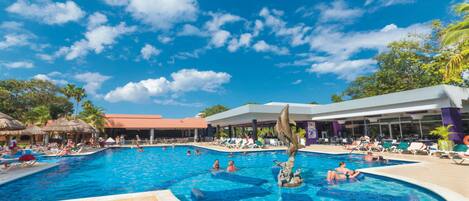 Una gran piscina al aire libre con varias pistas, rodeada de sillas de descanso y sombrillas, y un edificio con grandes ventanales al fondo.