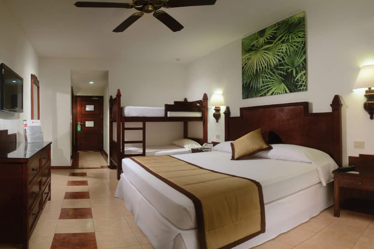 Una camera d'albergo con un letto a castello, una scrivania con televisione a schermo piatto, un letto con copriletto color beige, una lampada a parete e un quadro con motivi vegetali.
