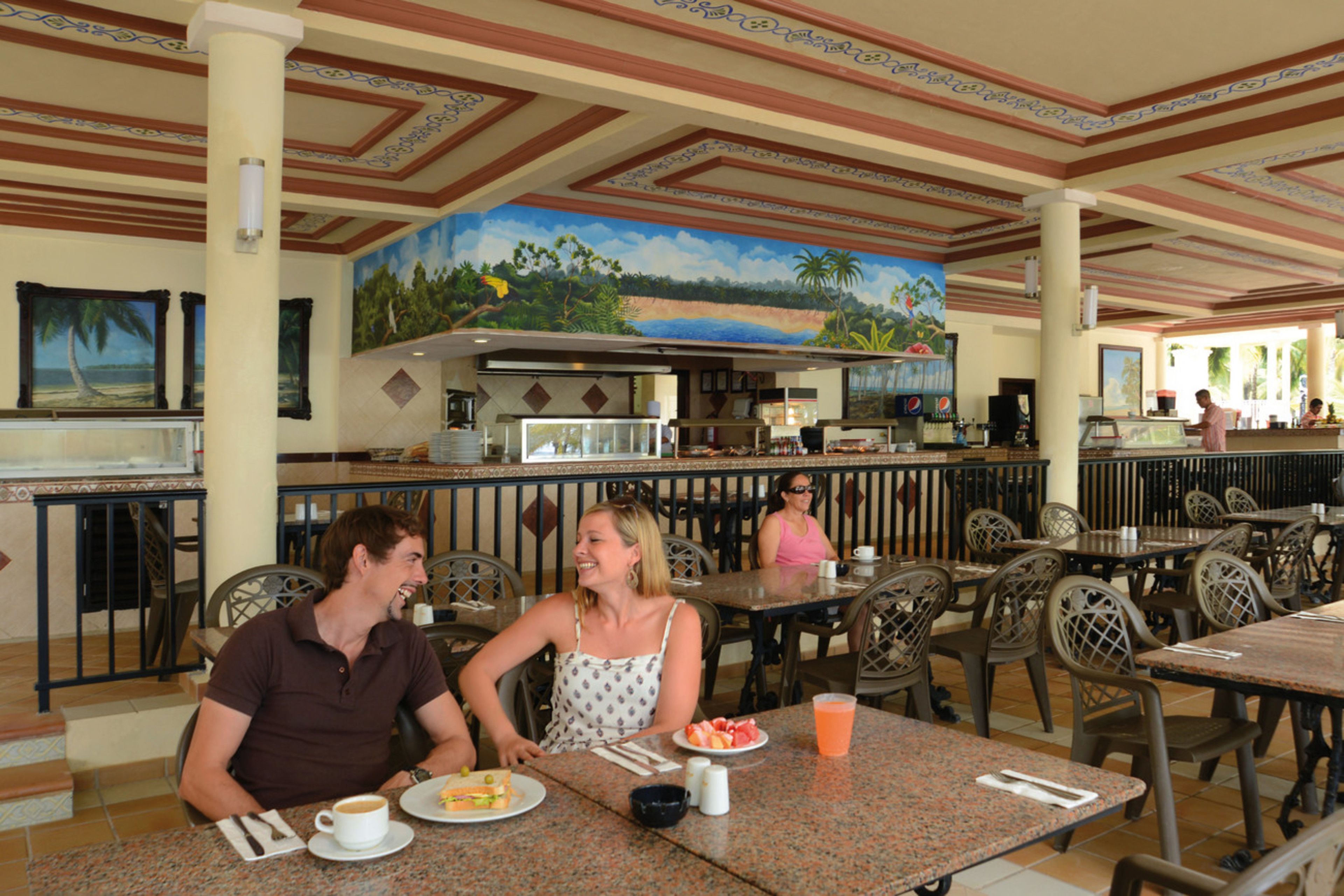 Un restaurante con un mural tropical, mesas al aire libre y una zona de bar.