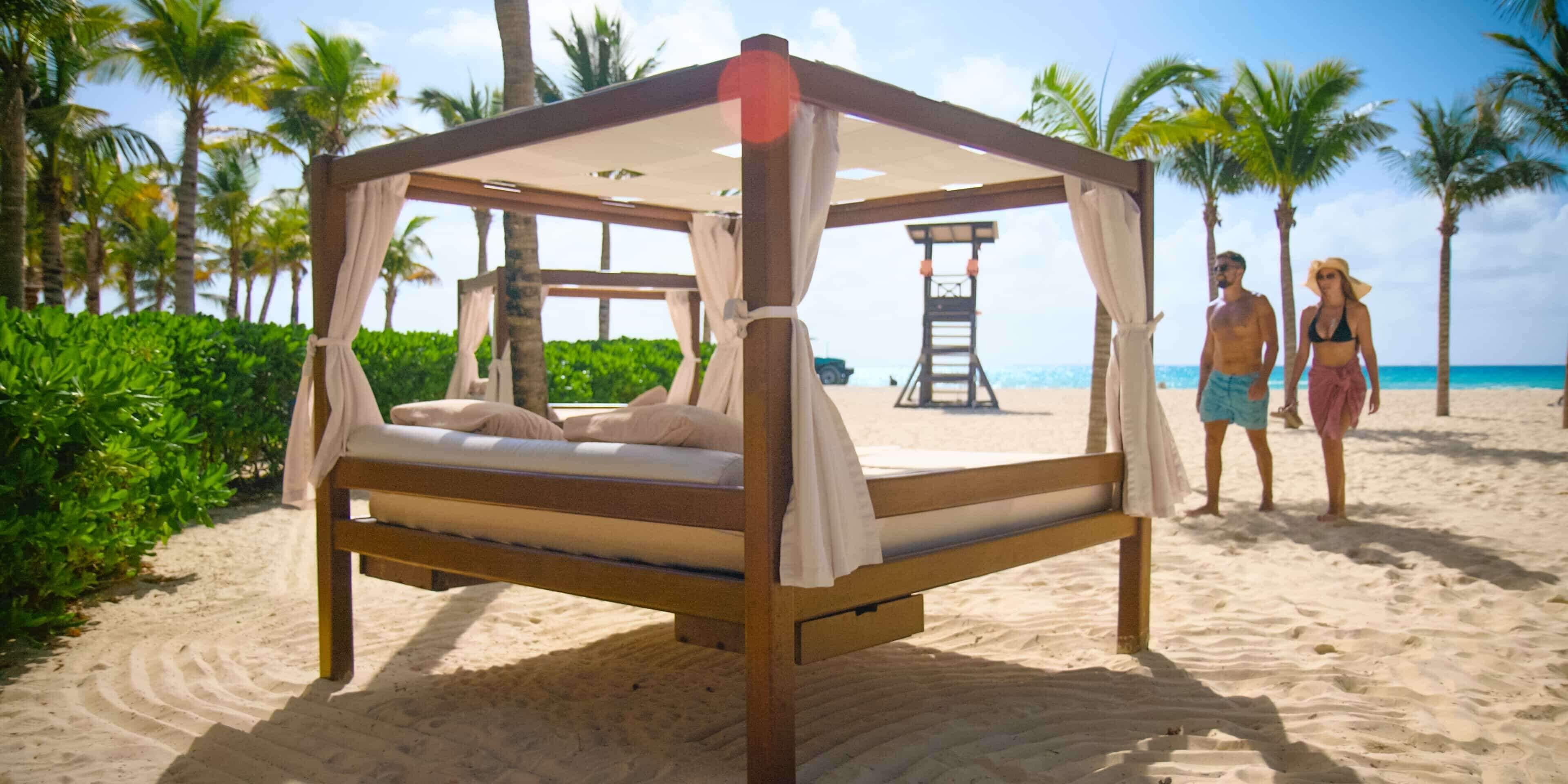 A bed with a canopy, a wooden bench, and a small table on a sandy beach with palm trees and a lifeguard tower in the background.