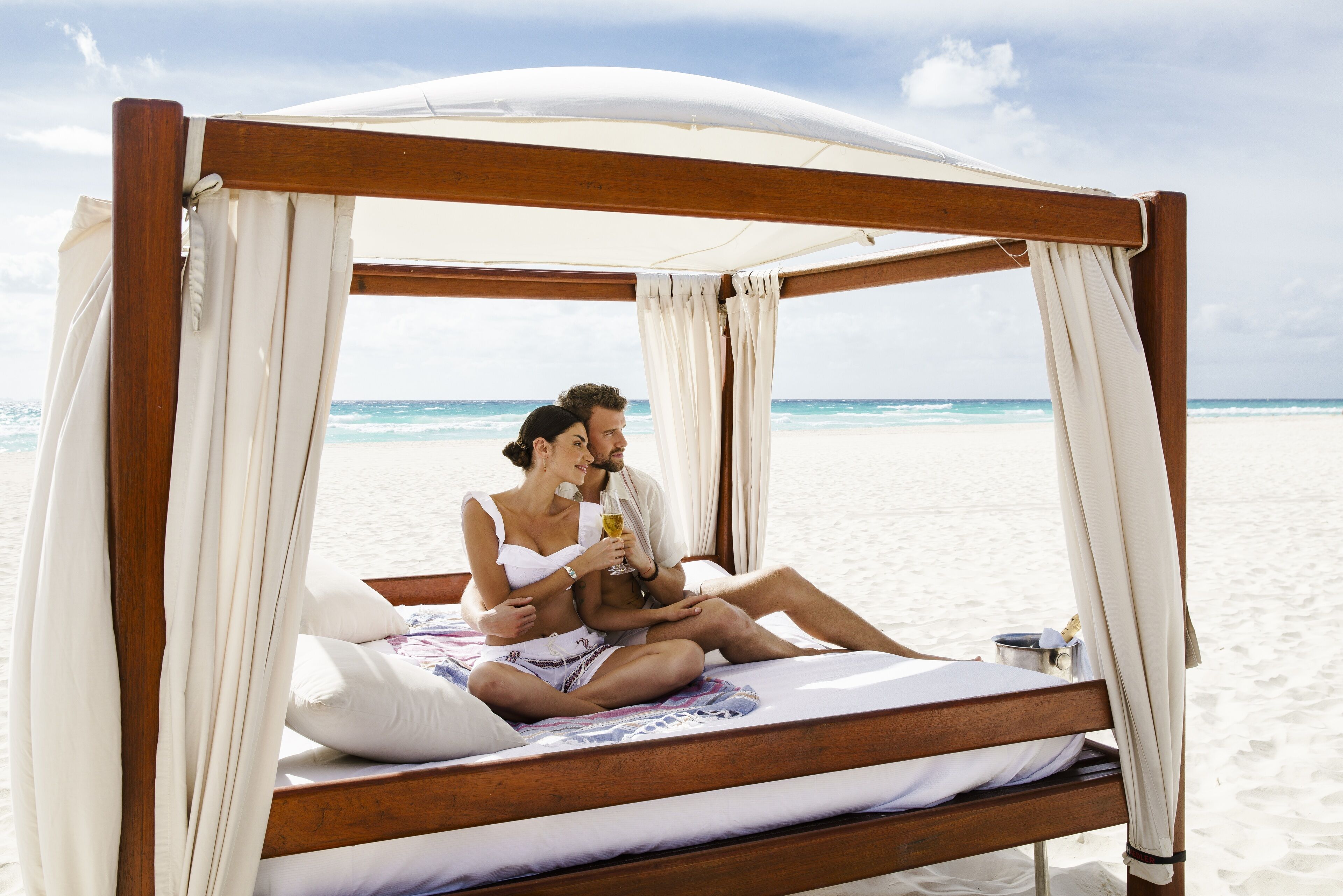 A wooden bed with a canopy, white bedding, and a view of the beach and ocean.