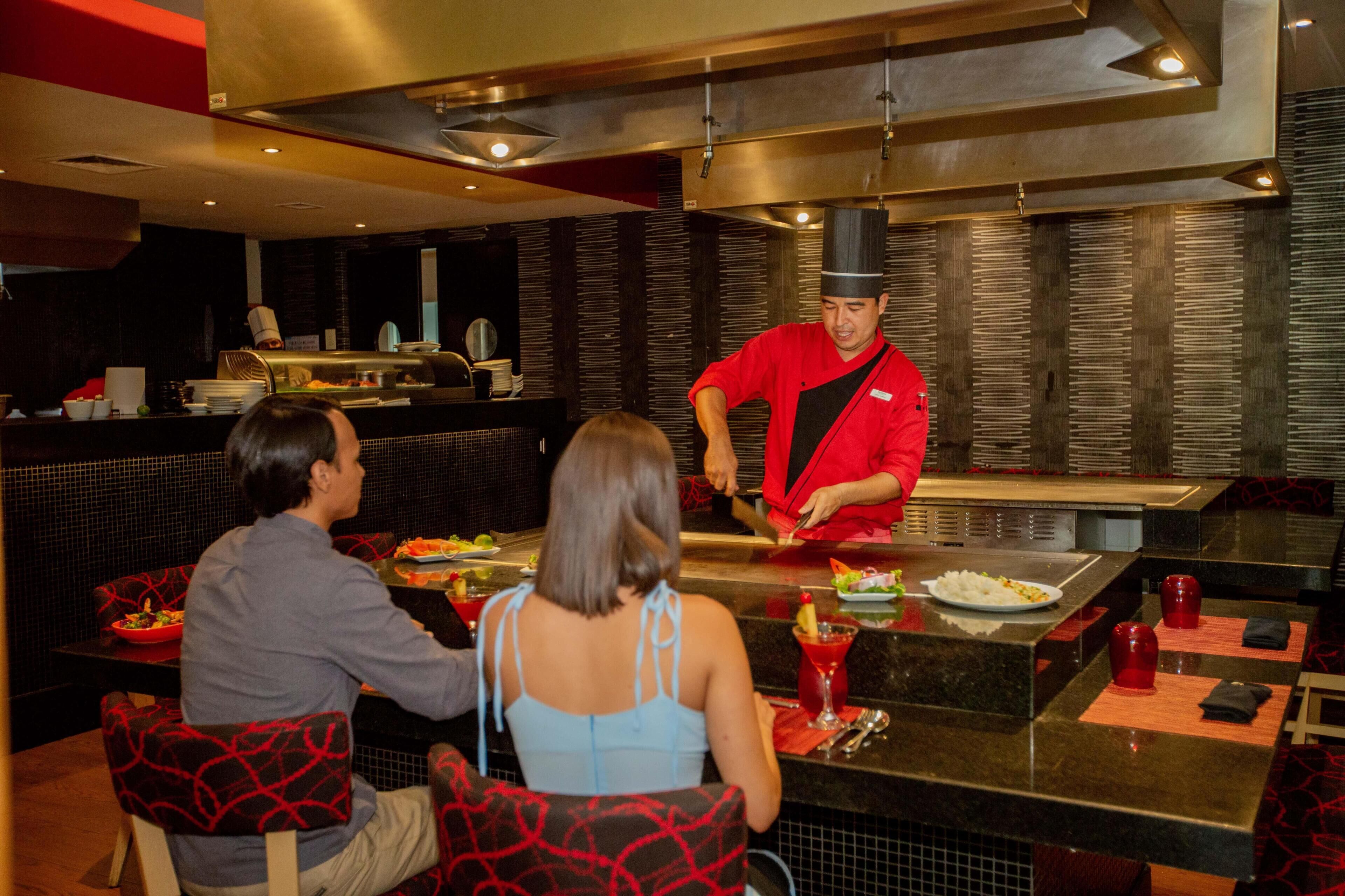 Un restaurante con un chef preparando comida en la barra, comensales sentados en mesas y una decoración de estilo moderno.