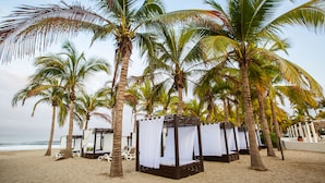 A beach with palm trees and a row of wooden beach huts with white curtains. - Occidental Nuevo Vallarta - All Inclusive (Nuevo Nayarit)