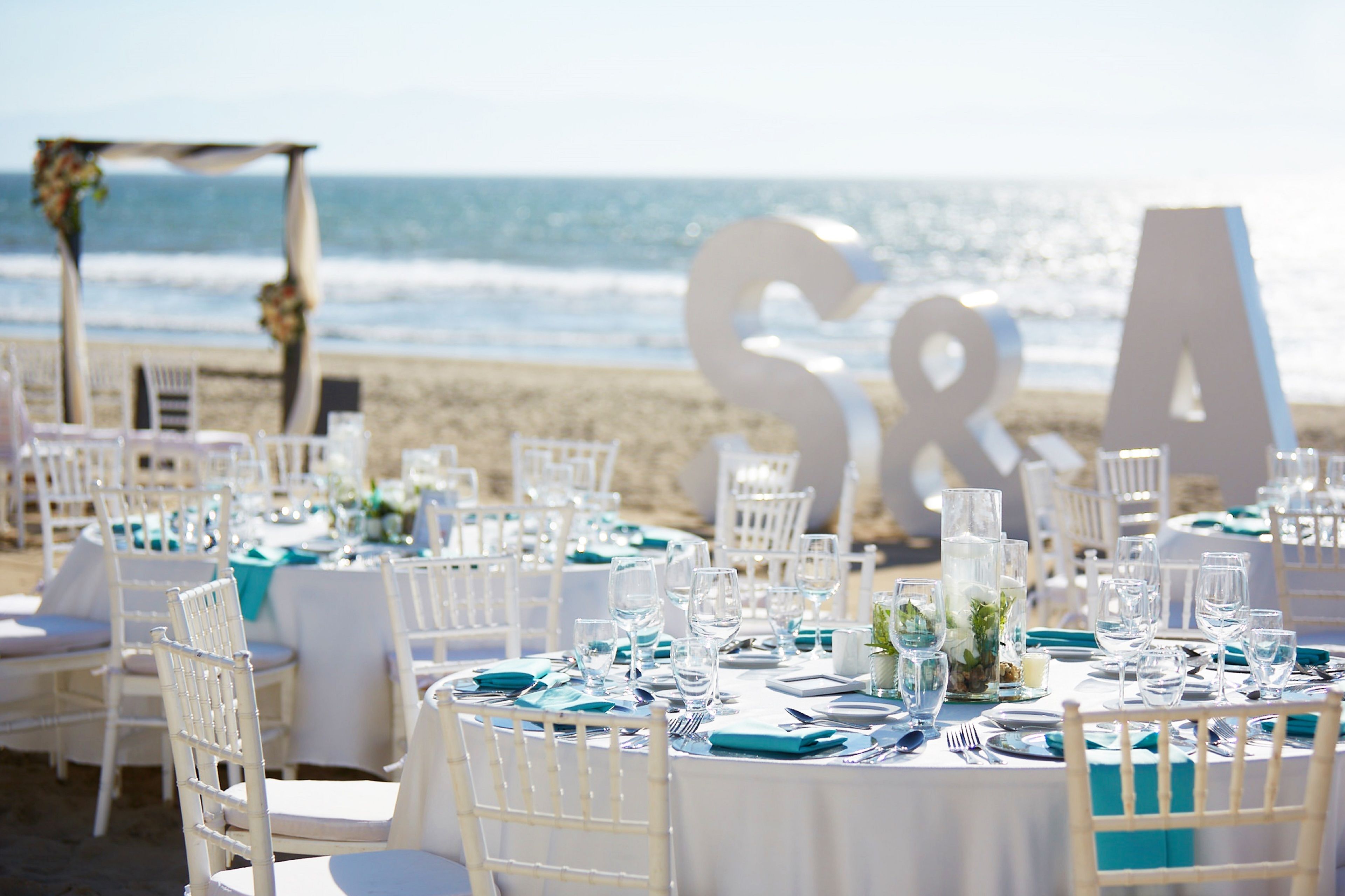A beachside wedding setup with white tables and chairs, large letters spelling "S & A" in the background, and a clear view of the ocean.