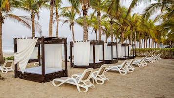 A beach with four wooden canopy beds and several white lounge chairs.