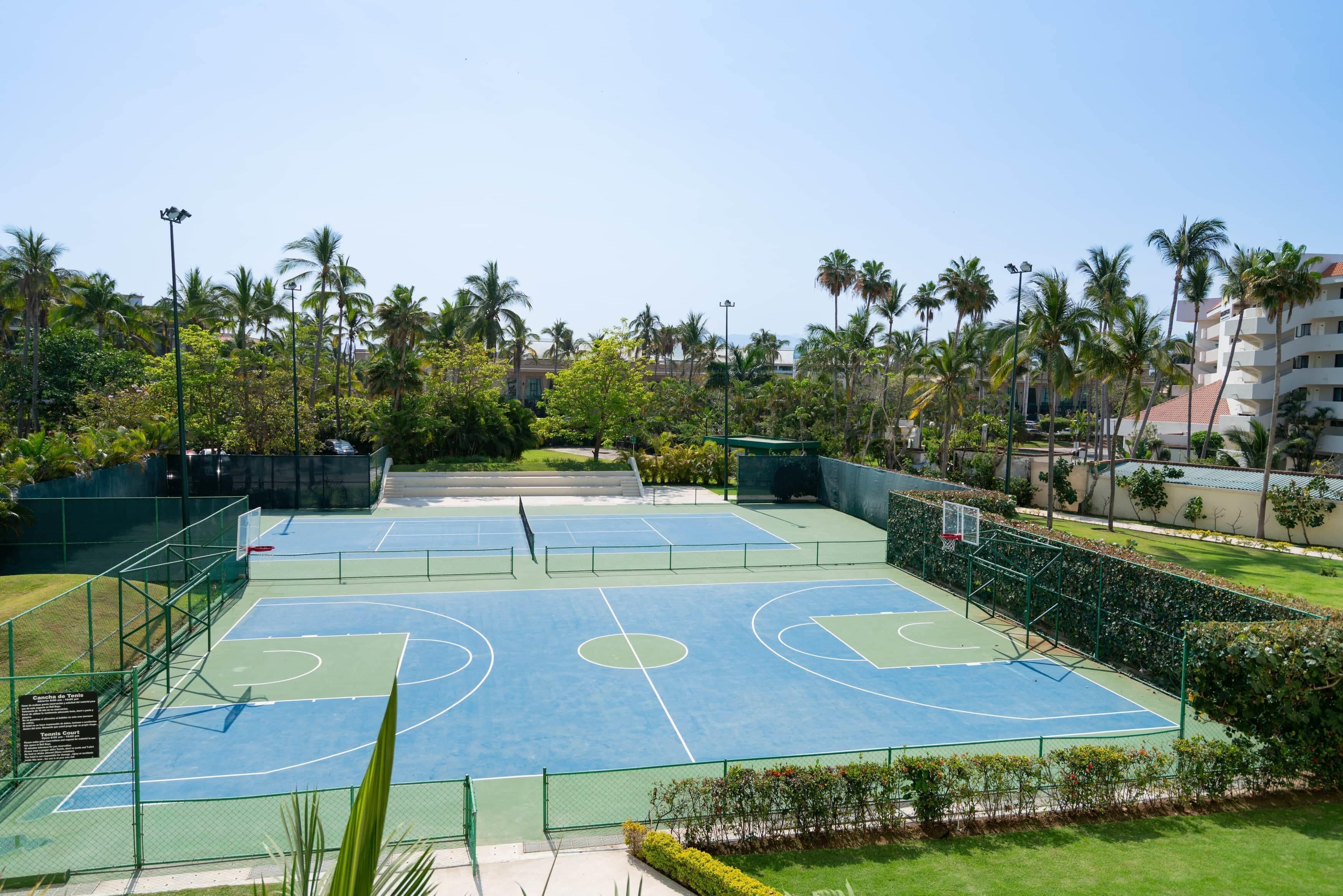 An outdoor basketball court with a green fence, surrounded by palm trees and a building in the background.