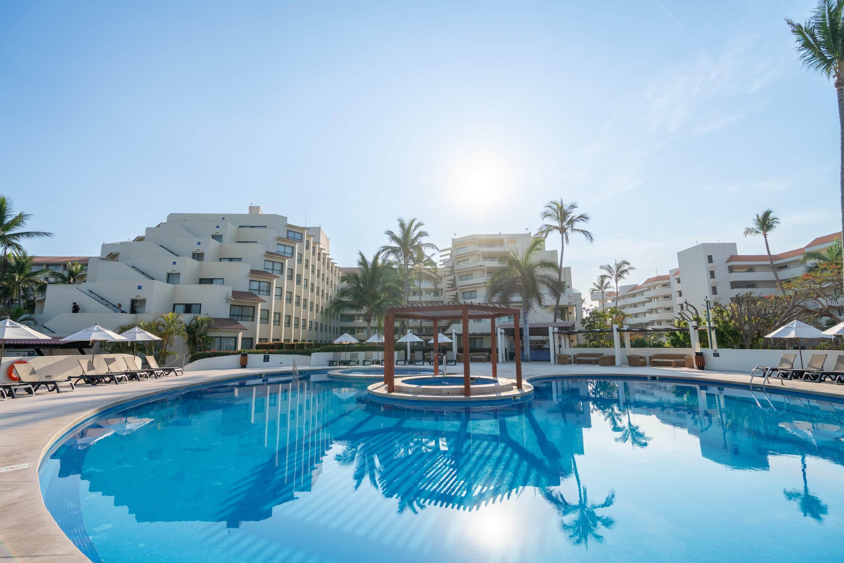 A swimming pool with lounge chairs and umbrellas, surrounded by palm trees and modern buildings.
