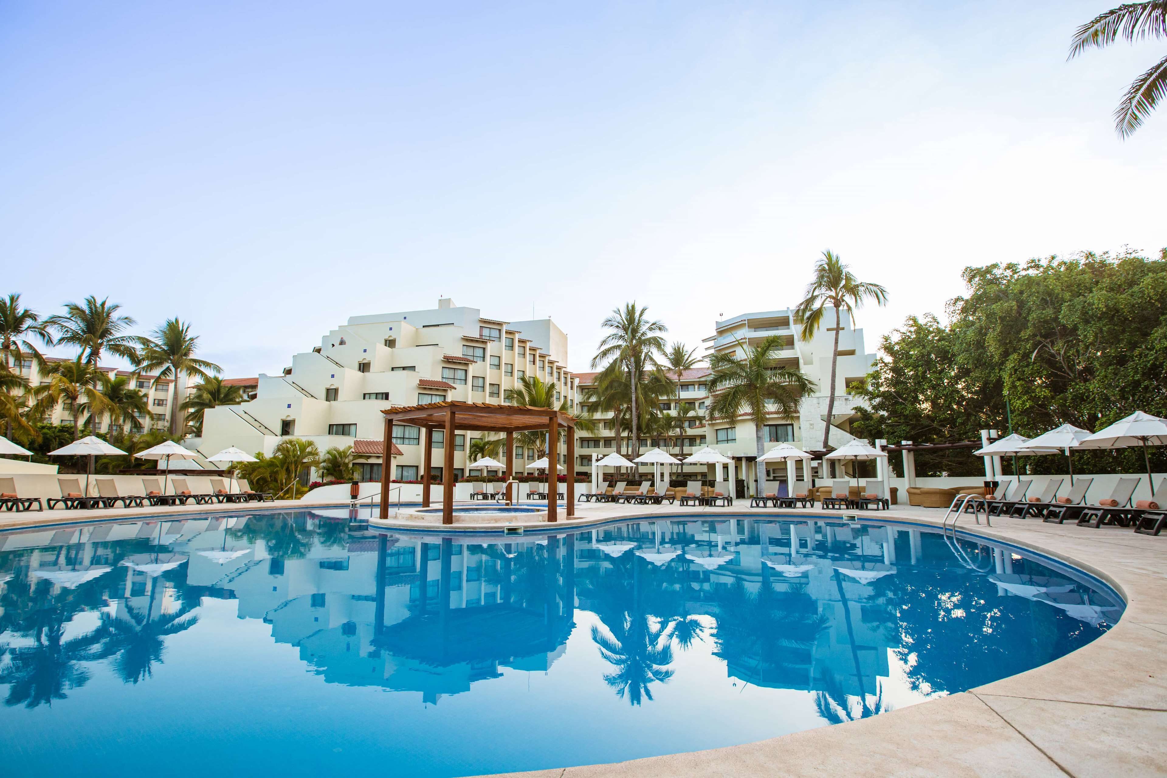Une piscine entourée de palmiers, avec des chaises longues et des parasols, et un immeuble à plusieurs étages doté de balcons.
