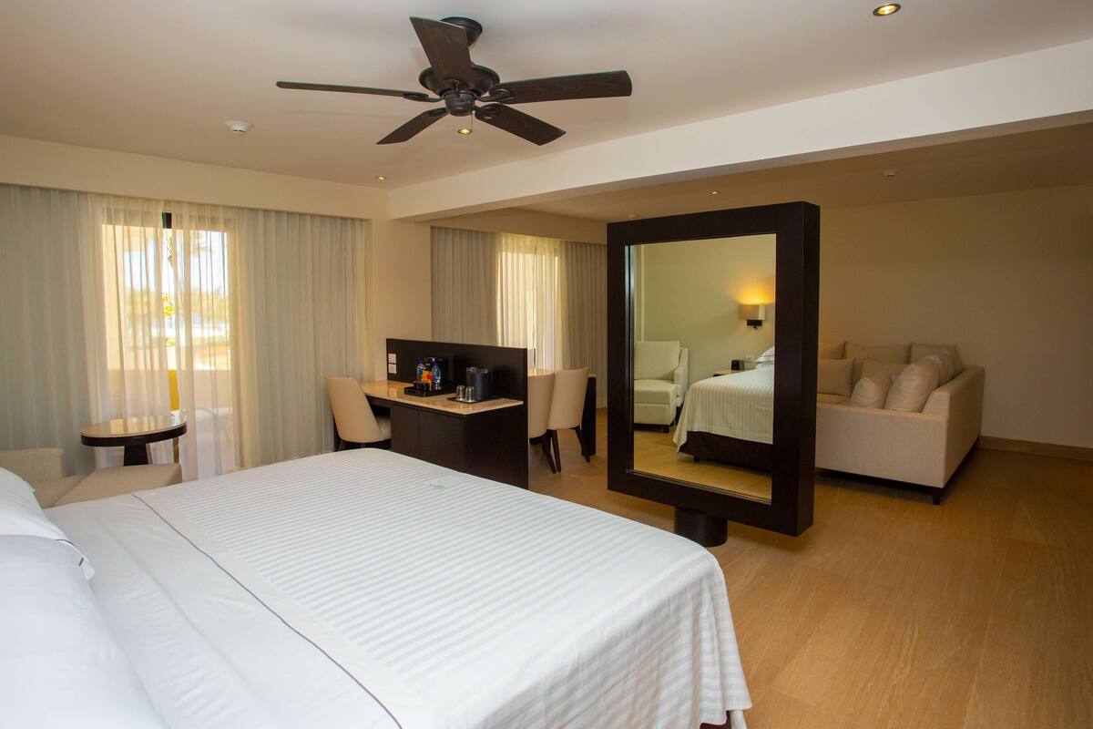 A hotel room with a large bed, a desk with a computer, a chair, a sofa, a ceiling fan, and a large mirror.