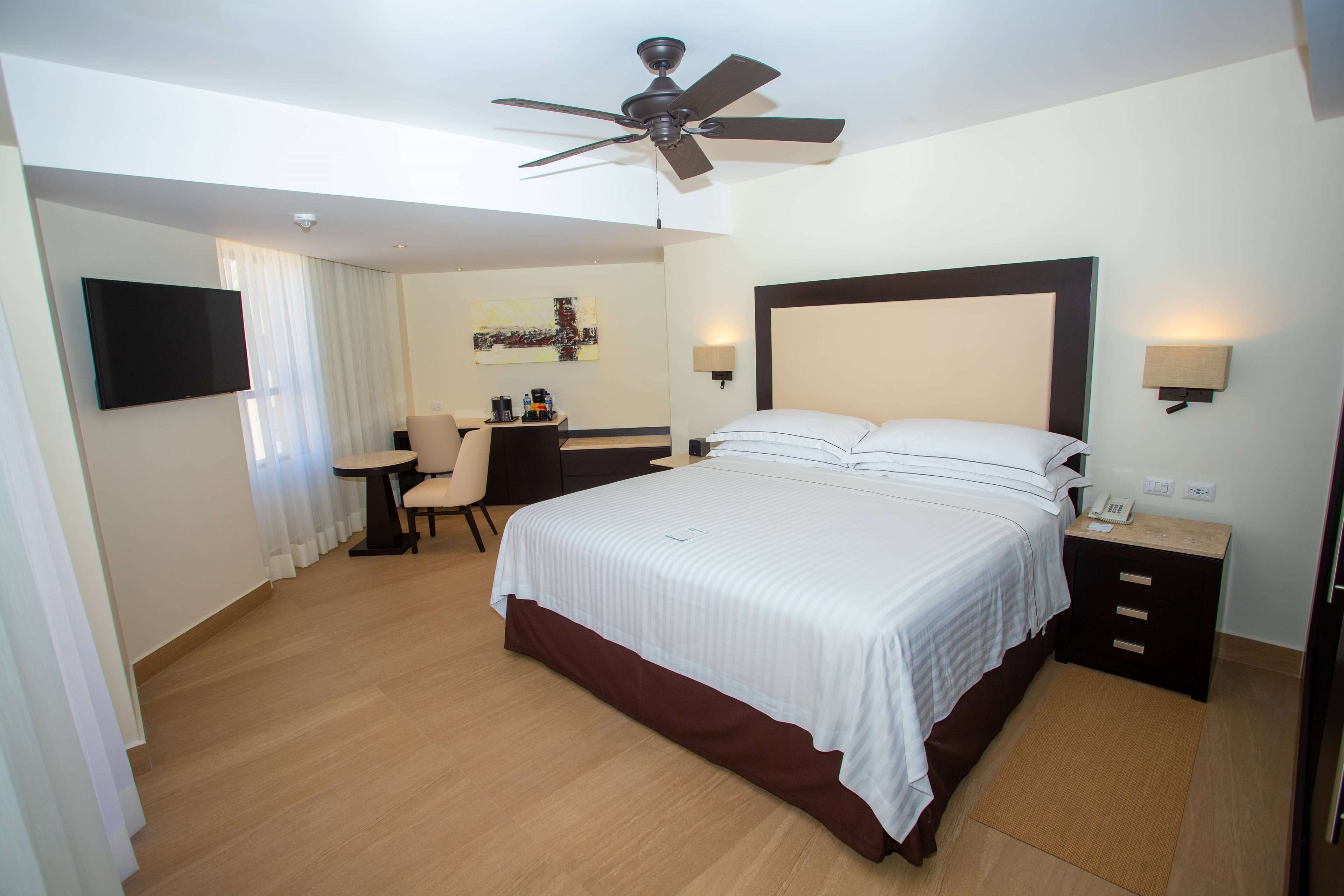 Habitación de hotel con cama, escritorio, silla, ventilador de techo y televisor.