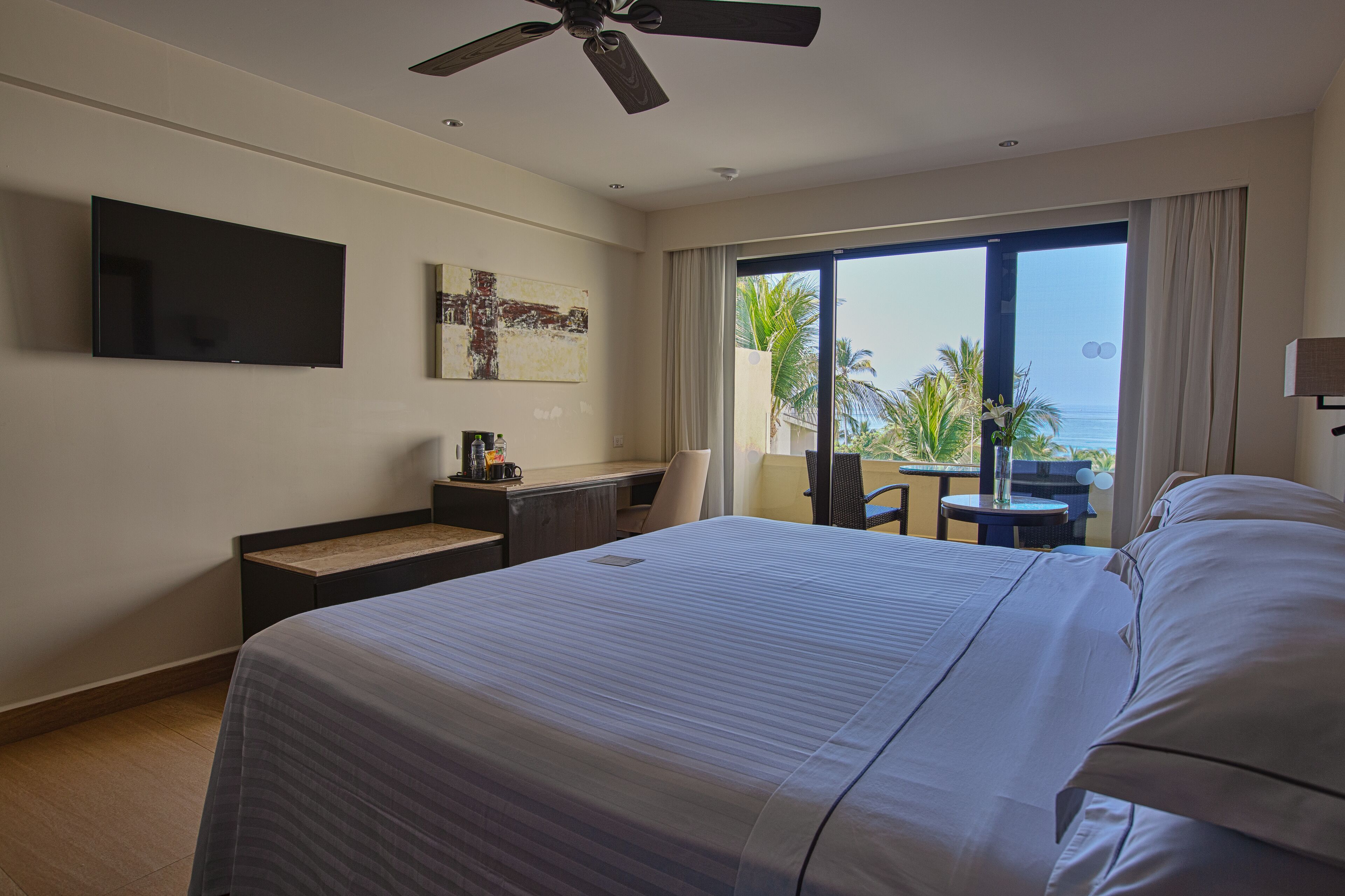 A hotel room with a large bed, a desk, a flat-screen TV, and a balcony with a view of the ocean.