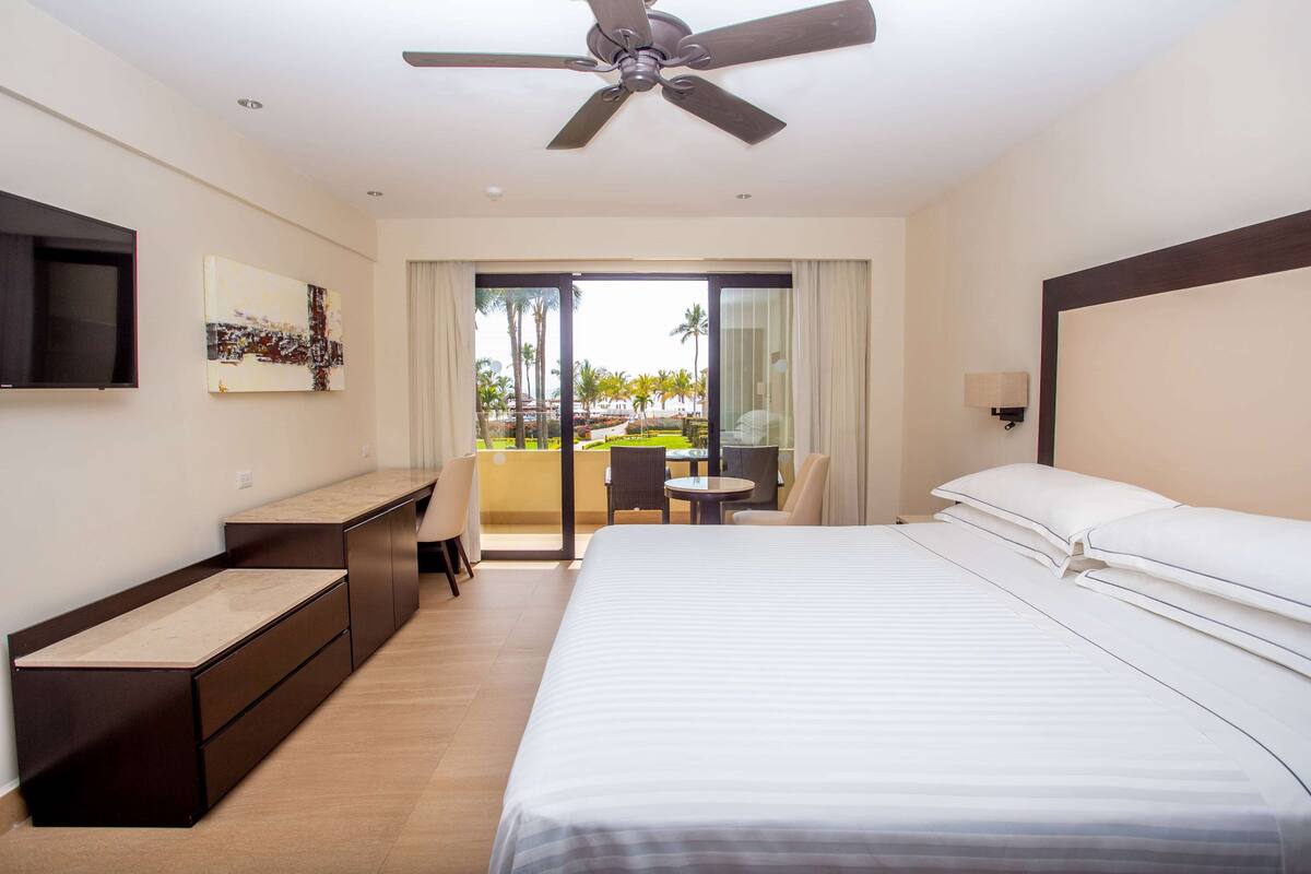 A hotel room with a bed, a desk, a TV, a ceiling fan, and a balcony with a view of palm trees.