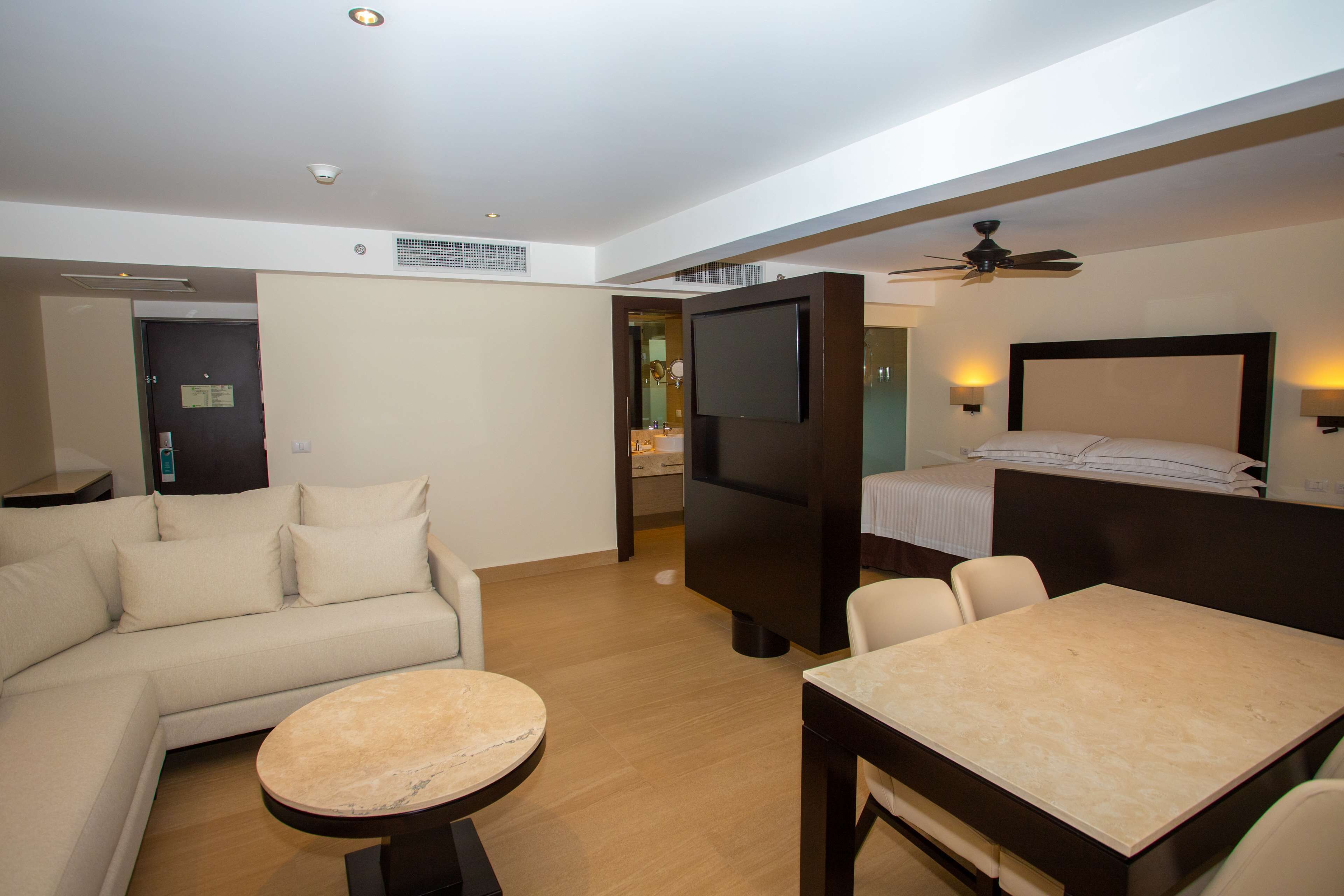A modern hotel room with a sofa, a round table, a bed with a headboard, a television, and a ceiling fan.