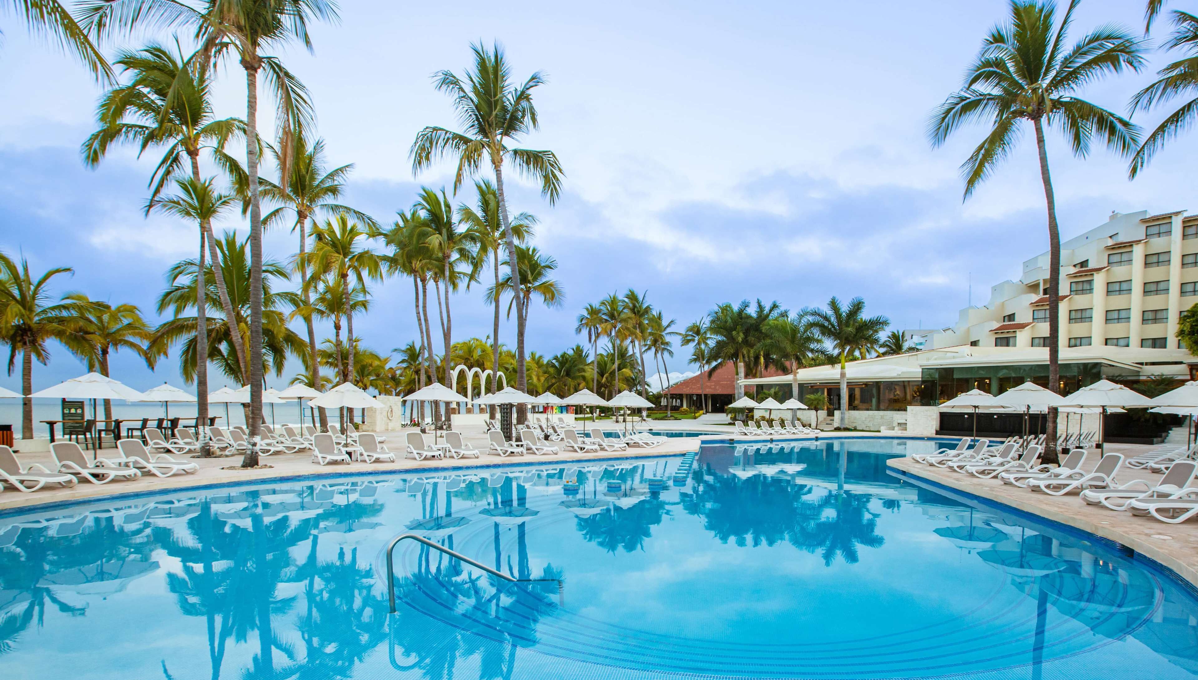 A pool area with lounge chairs and umbrellas, surrounded by palm trees and a hotel building.