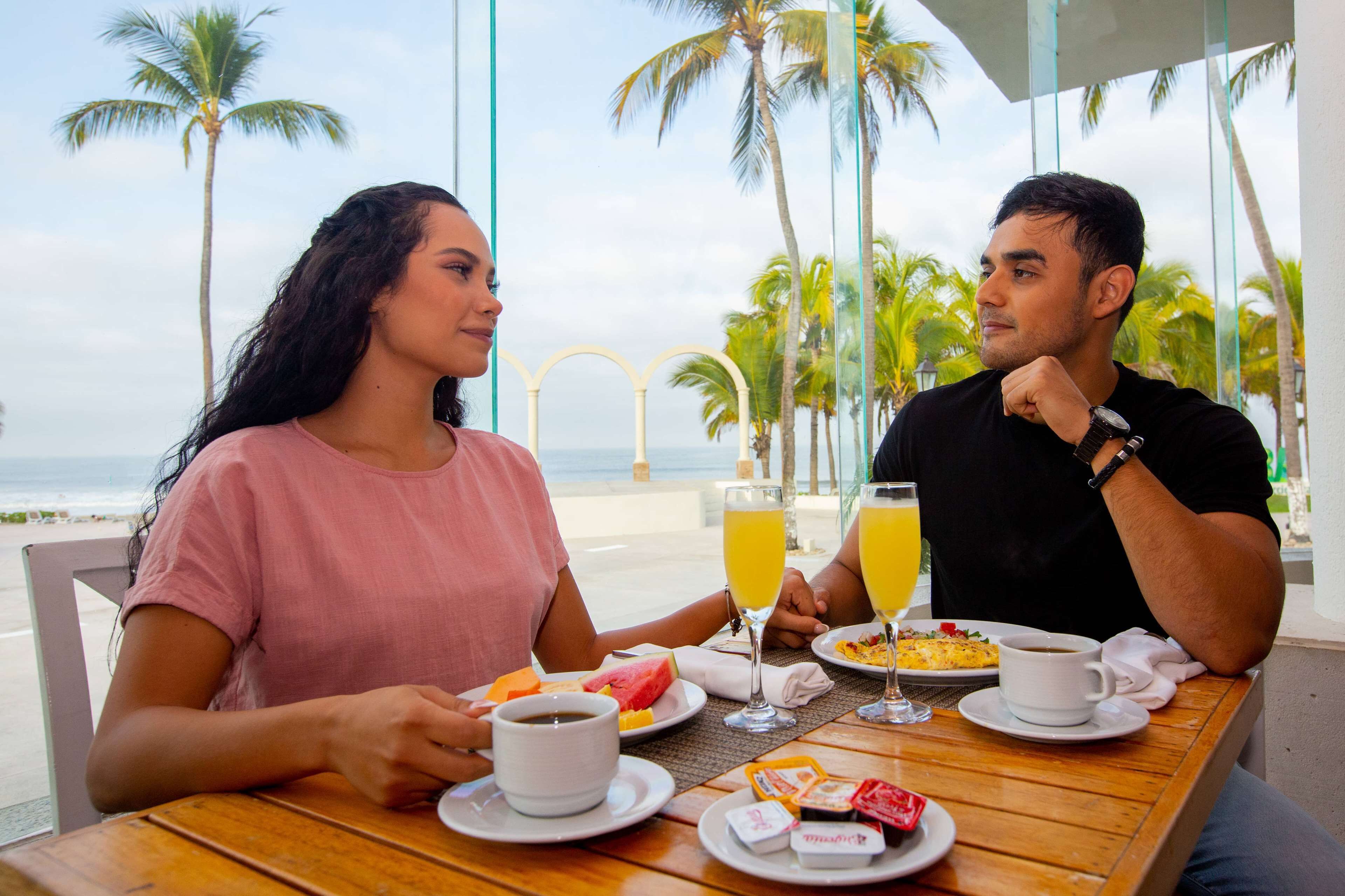 Una pareja cenando en una mesa al aire libre con vista a la playa y palmeras.
