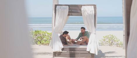 A wooden gazebo with white curtains on a beach.