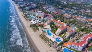 An aerial view of a coastal resort with multiple buildings, a beach, and several swimming pools.