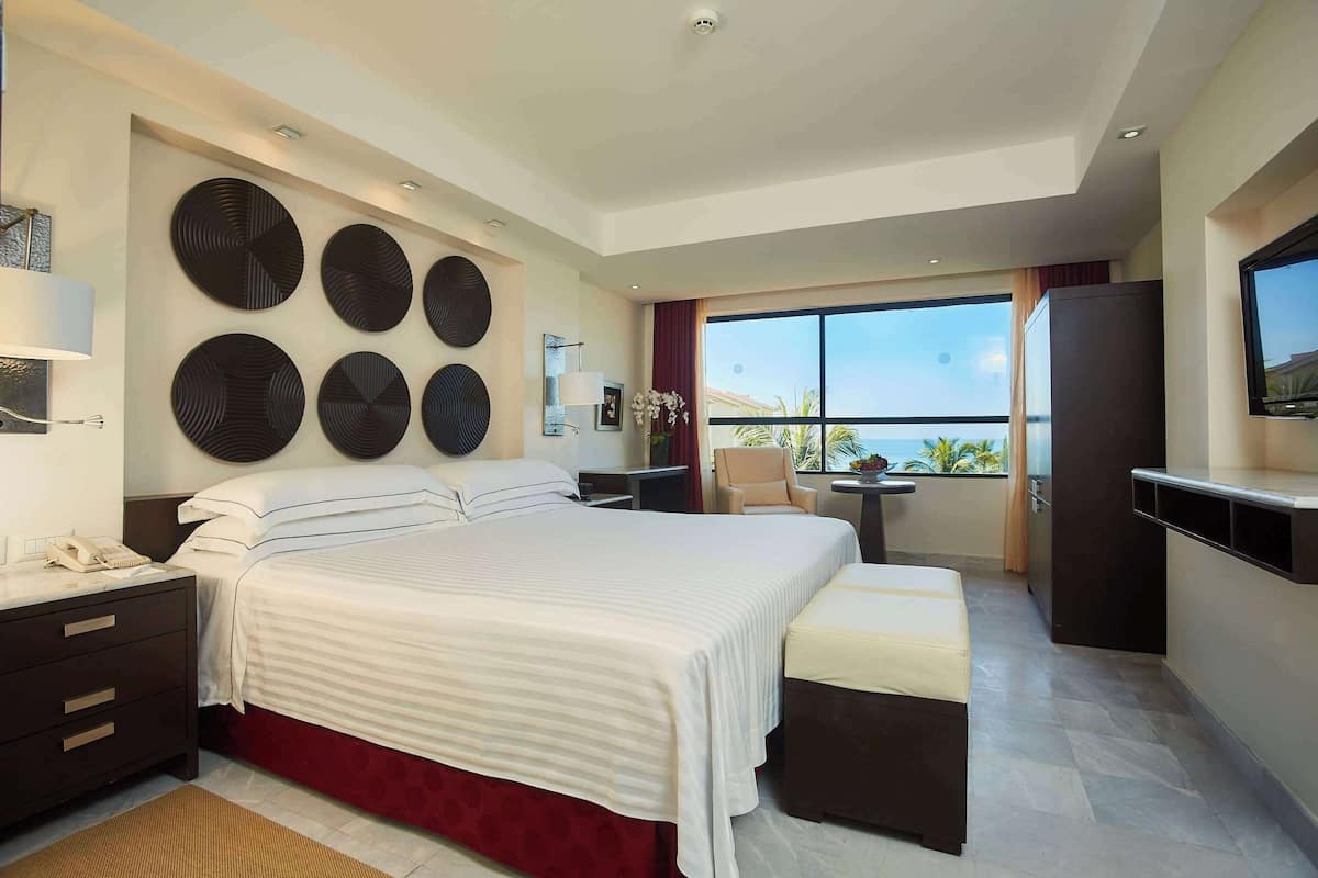 A modern hotel room with a large bed, a desk, a chair, a TV, and a balcony with a view.