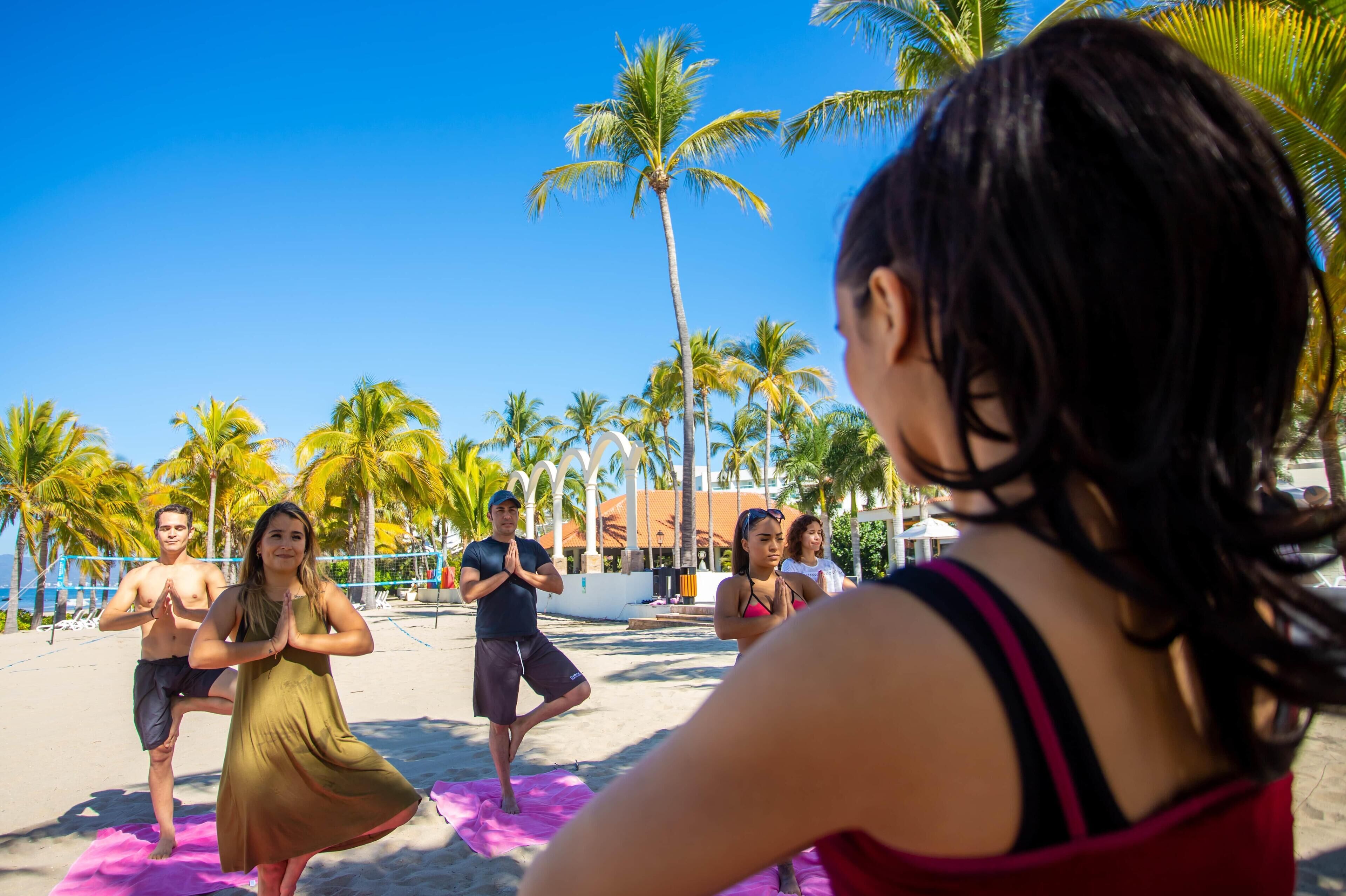 A group of people practicing yoga on a beach with palm trees and a clear sky in the background.