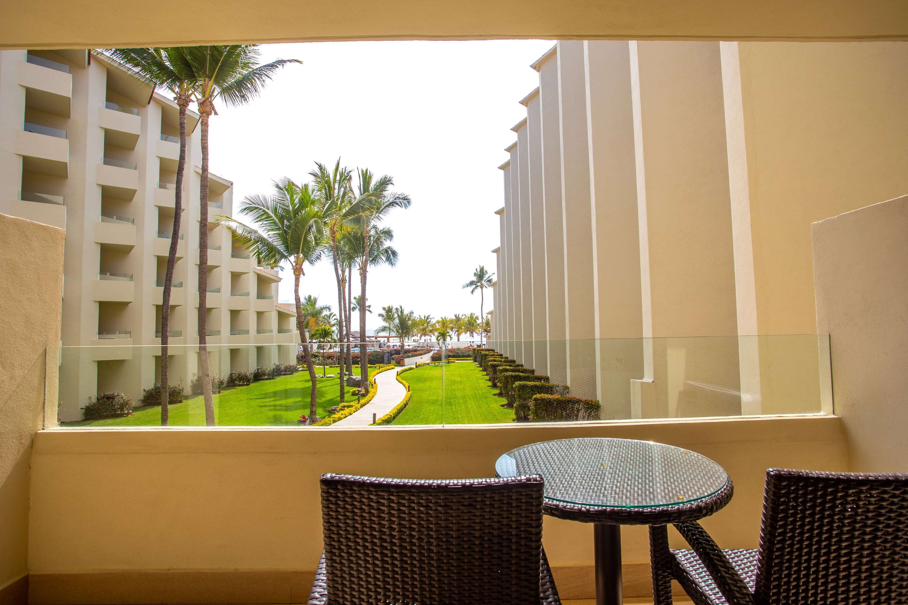 A balcony with wicker chairs and a glass table, overlooking a well-maintained lawn and palm trees, with a modern building in the background.